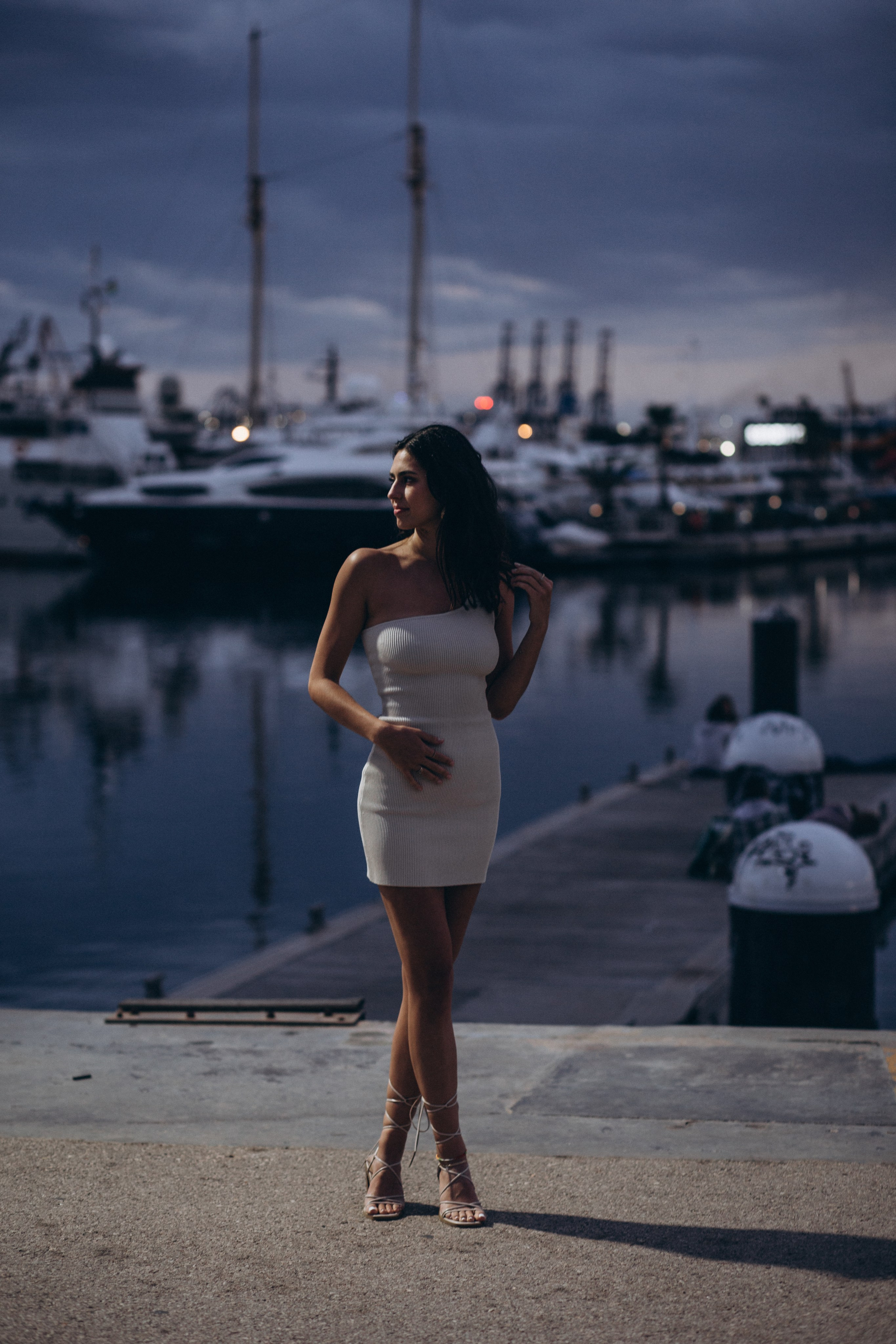 Элегантная женщина в белом платье позирует у марины на закате на фоне роскошных яхт в Валенсии, Испания — стильная и современная портретная фотография, передающая прибрежный гламур, мягкий вечерний свет и средиземноморскую атмосферу, идеально подходит для портретных фотосессий в Валенсии и Испании.