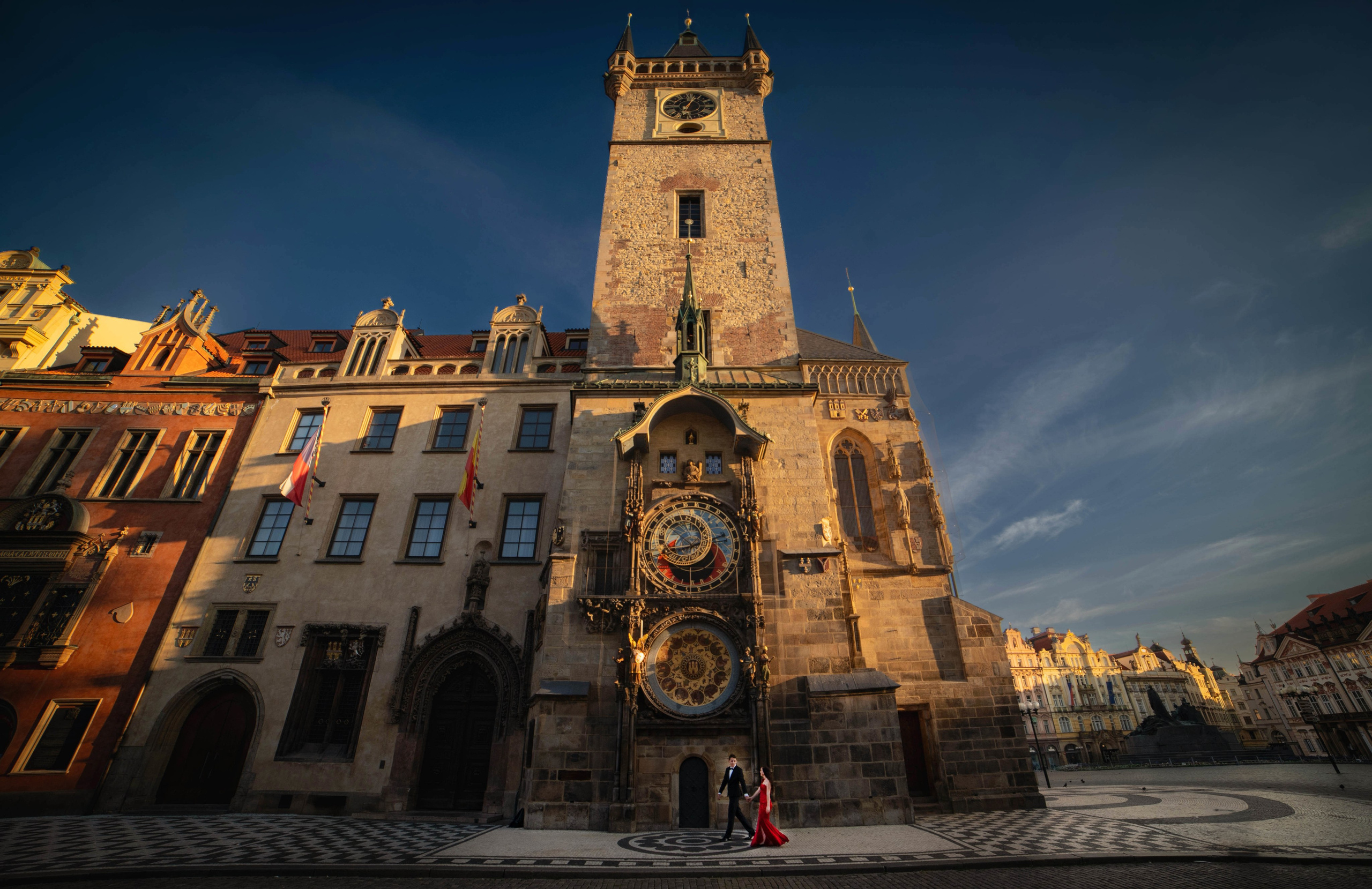 As the medieval tower above Prague's Astronomical Clock towers above, a stylishly dressed woman in red evening gown walks hand in hand with her tuxedo wearing partner underneath all alone