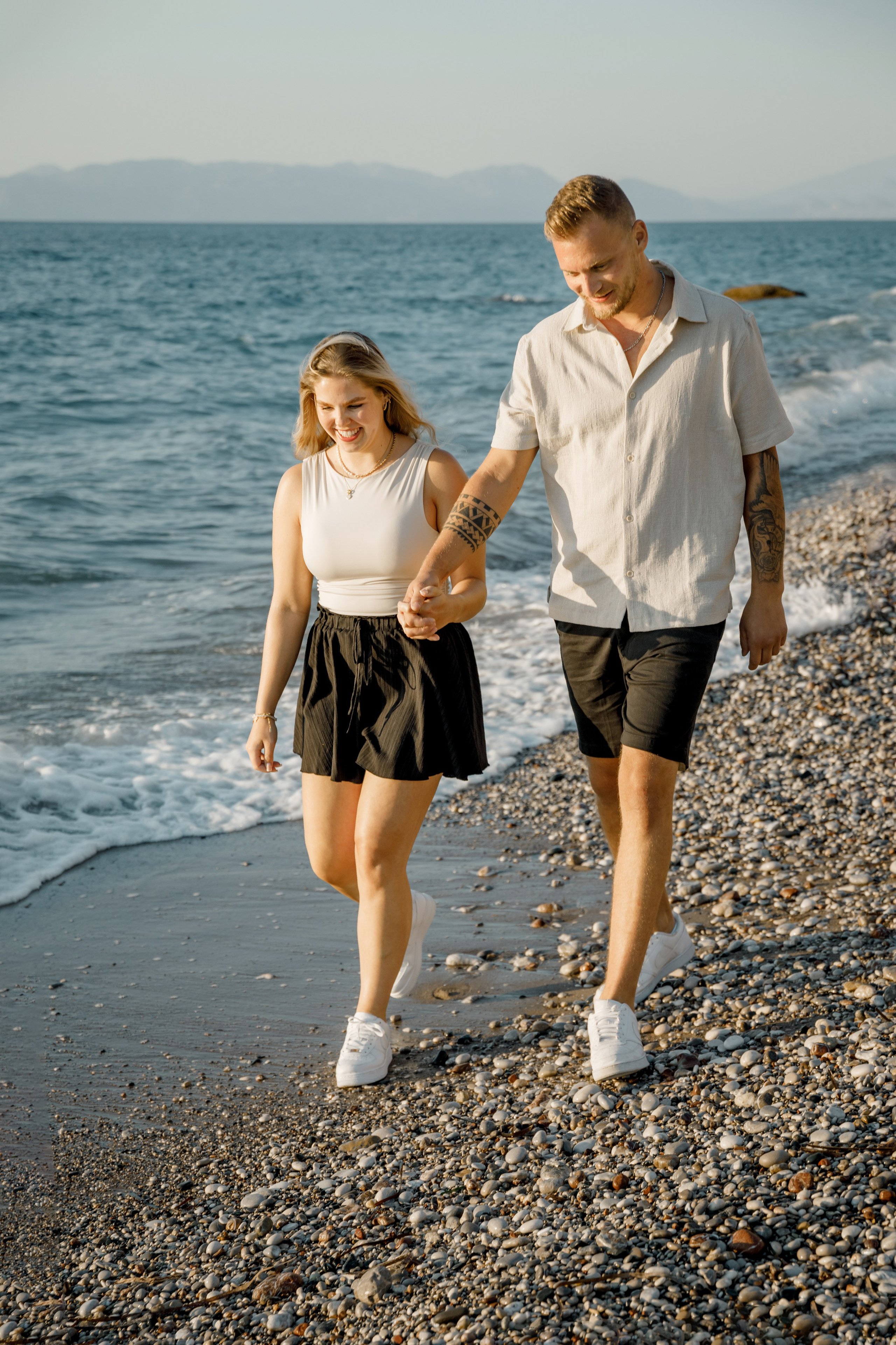 "Ein Paar spaziert Hand in Hand am steinigen Ufer von Kato Petres auf Rhodos entlang. Sie gehen direkt am Wasser, während im Hintergrund die hohen, zerklüfteten Kalksteinfelsen aufragen und das blaue Meer sanft gegen die Kiesel brandet."