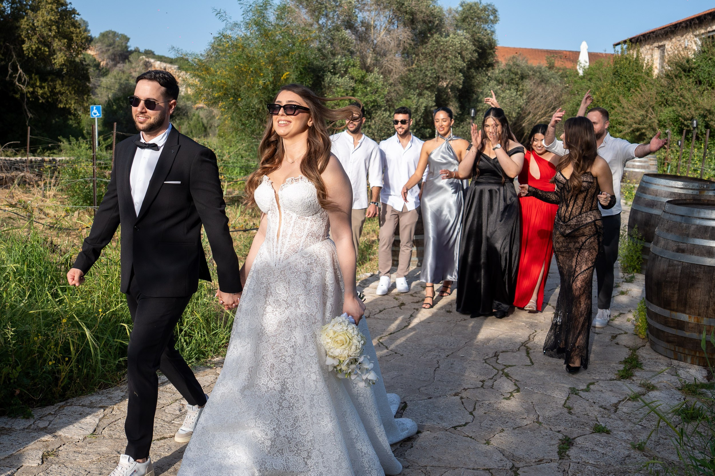 A bride and groom walk outdoors with their wedding guests behind them