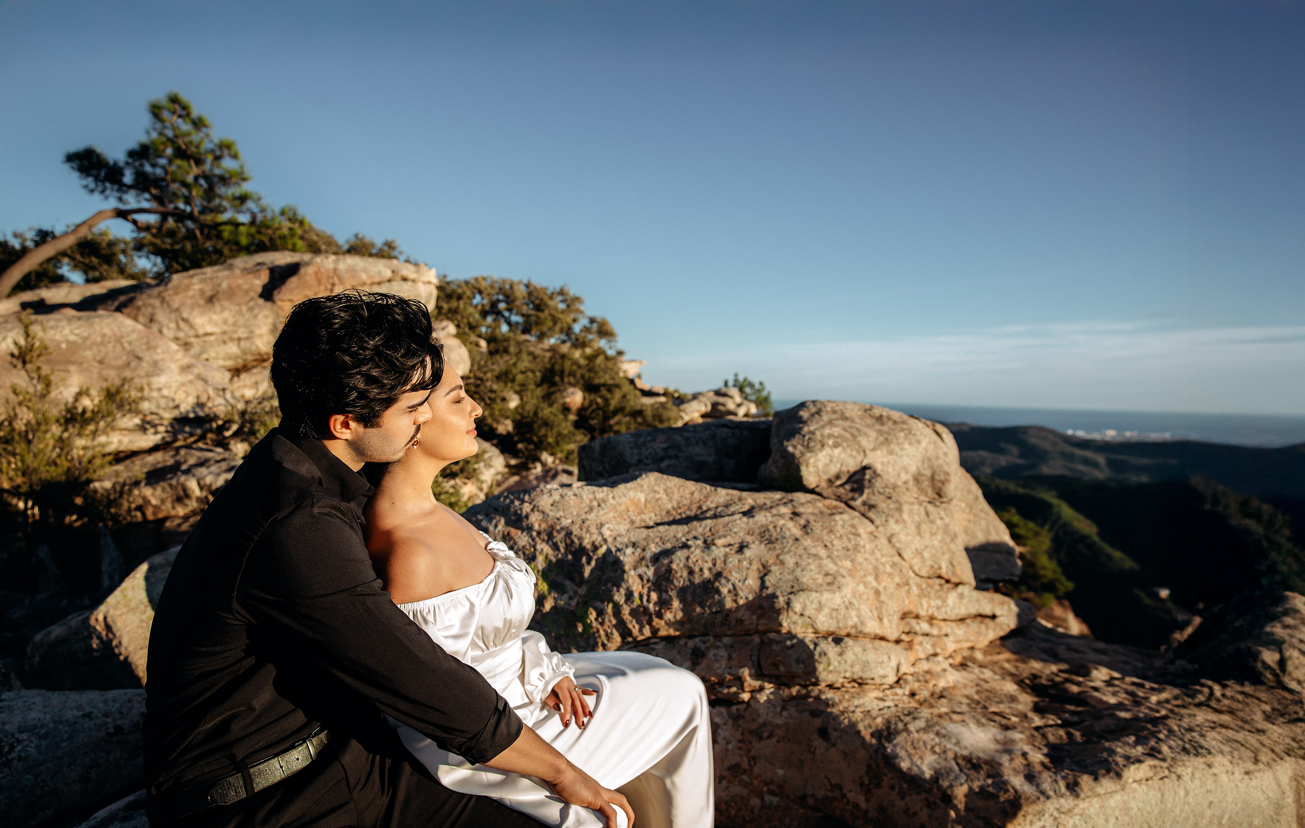 Groom sitting beside the bride on a mountain viewpoint during an intimate elopement in Barcelona, Spain. This relaxed destination wedding portrait captures quiet romance surrounded by dramatic natural scenery.