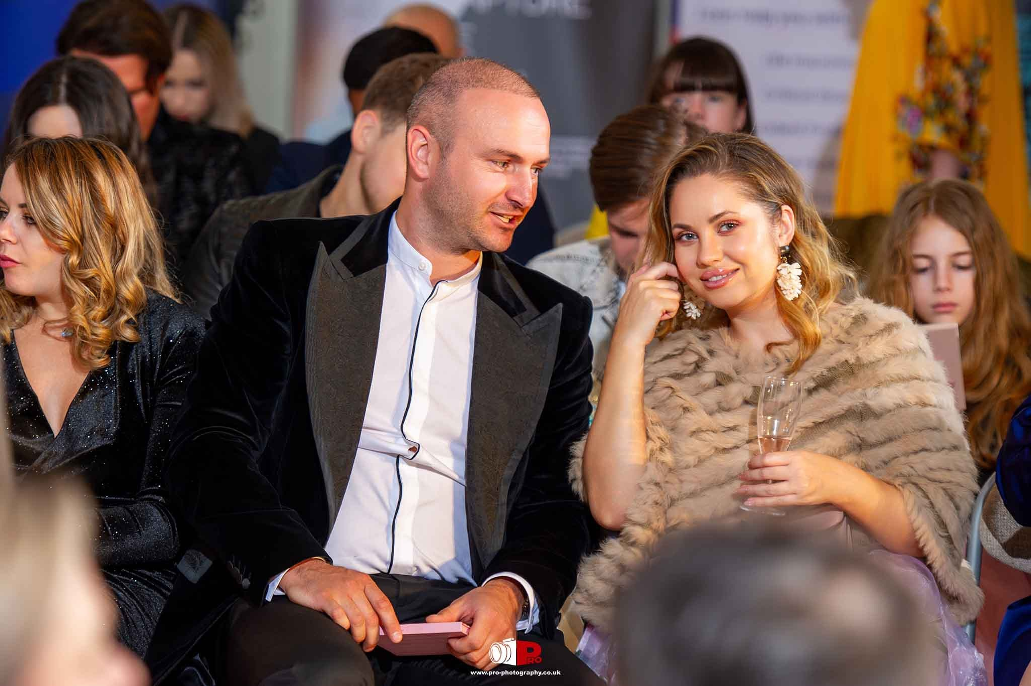 A smiling man and woman share a lighthearted moment with champagne glasses at a social gathering.