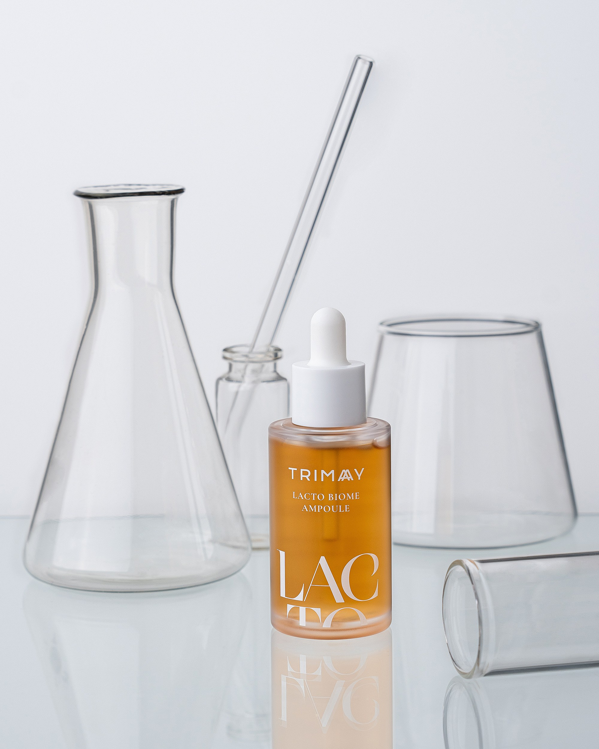 Trimay Lacto Biome Ampoule damlalıq şüşəsi, şəffaf laboratoriya kolbaları və şüşə qablar fonunda təqdimat.