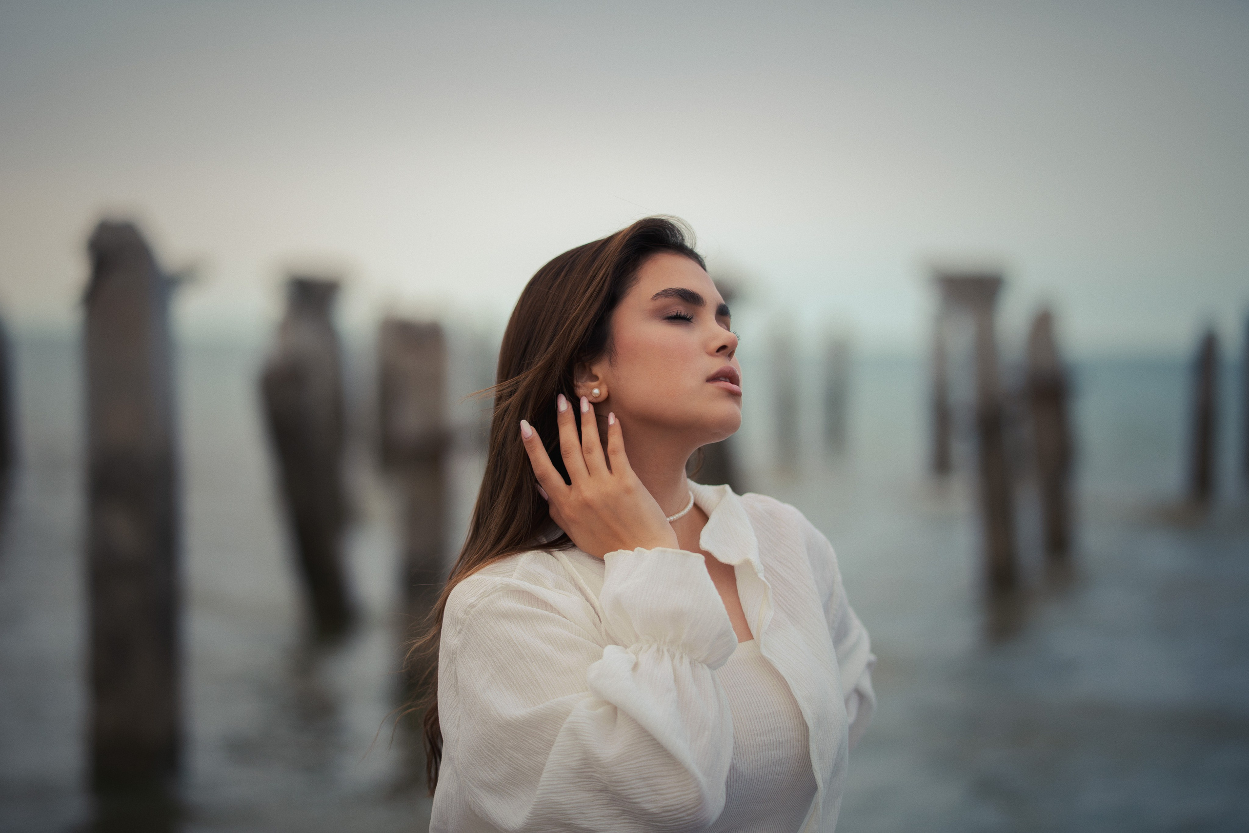 Female portrait photography by Mehran Hadad featuring a young woman posing beside weathered wooden posts at the seaside during golden hour with soft natural light.
