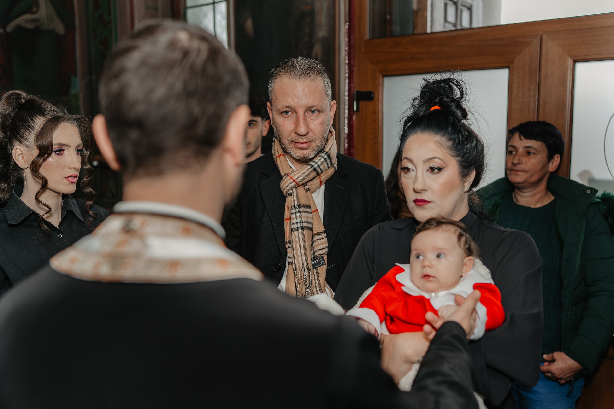 Începutul ceremoniei de botez a fetiței Eveline Maria în Biserica Grivița din Vaslui. Nașul ține pruncul în brațe în fața altarului, în timp ce preotul pregătește sfânta taină. În cadru se vede și mama plină de emoție. Fotografie naturală de botez.