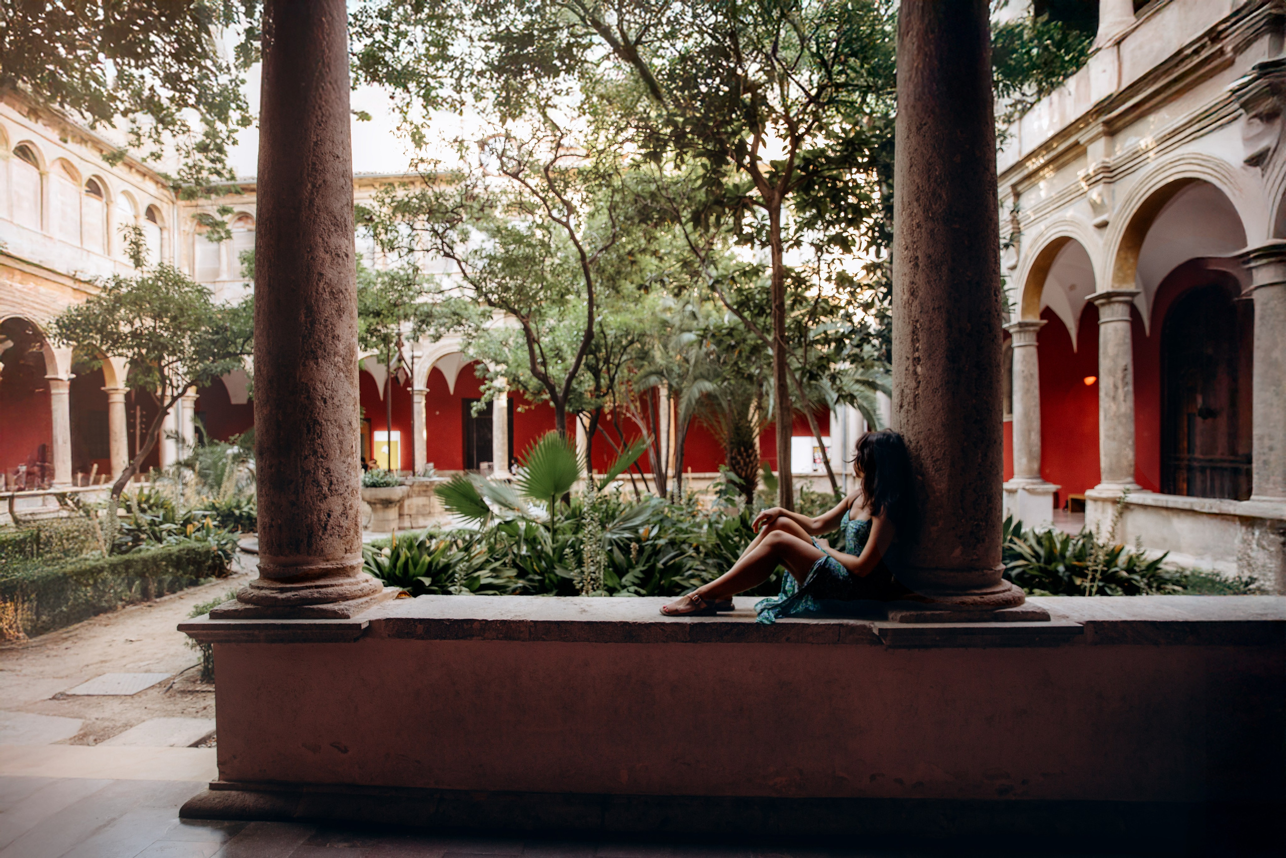 Женская фотосессия в Валенсии, Испания, где женщина изящно сидит среди исторических каменных колонн и арок в зелёном средиземноморском дворике, окружённая растительностью и мягким естественным светом — идеально для женской портретной фотографии, лайфстайл-съёмок и элегантных женских фотосессий в Валенсии и по всей Испании.
