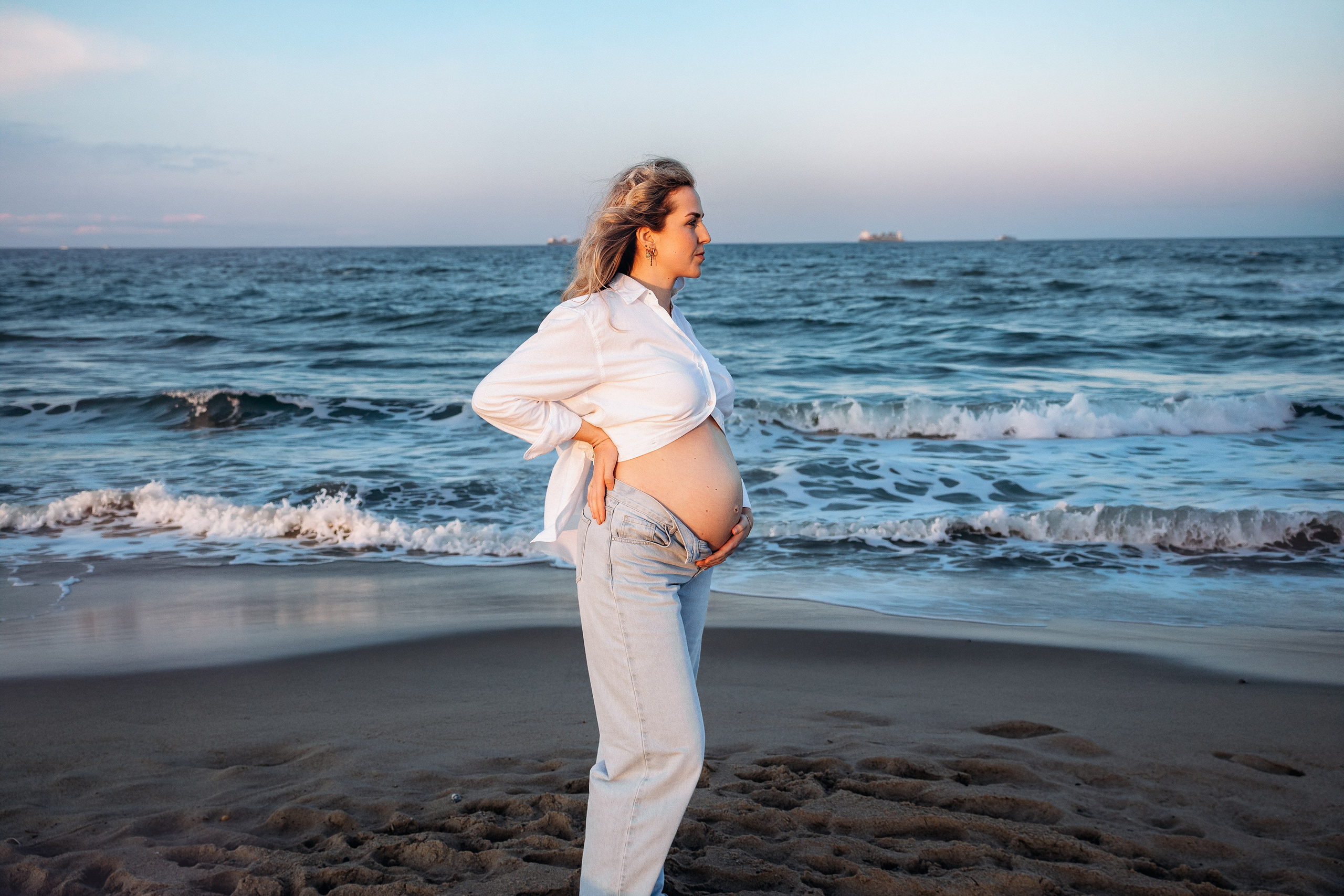 Sesión de fotos de embarazo en Valencia, España, con una mujer embarazada de pie junto al mar con un conjunto blanco, sosteniendo suavemente su barriga, capturada con suave luz natural de playa — ideal para sesiones maternity en Valencia y España.