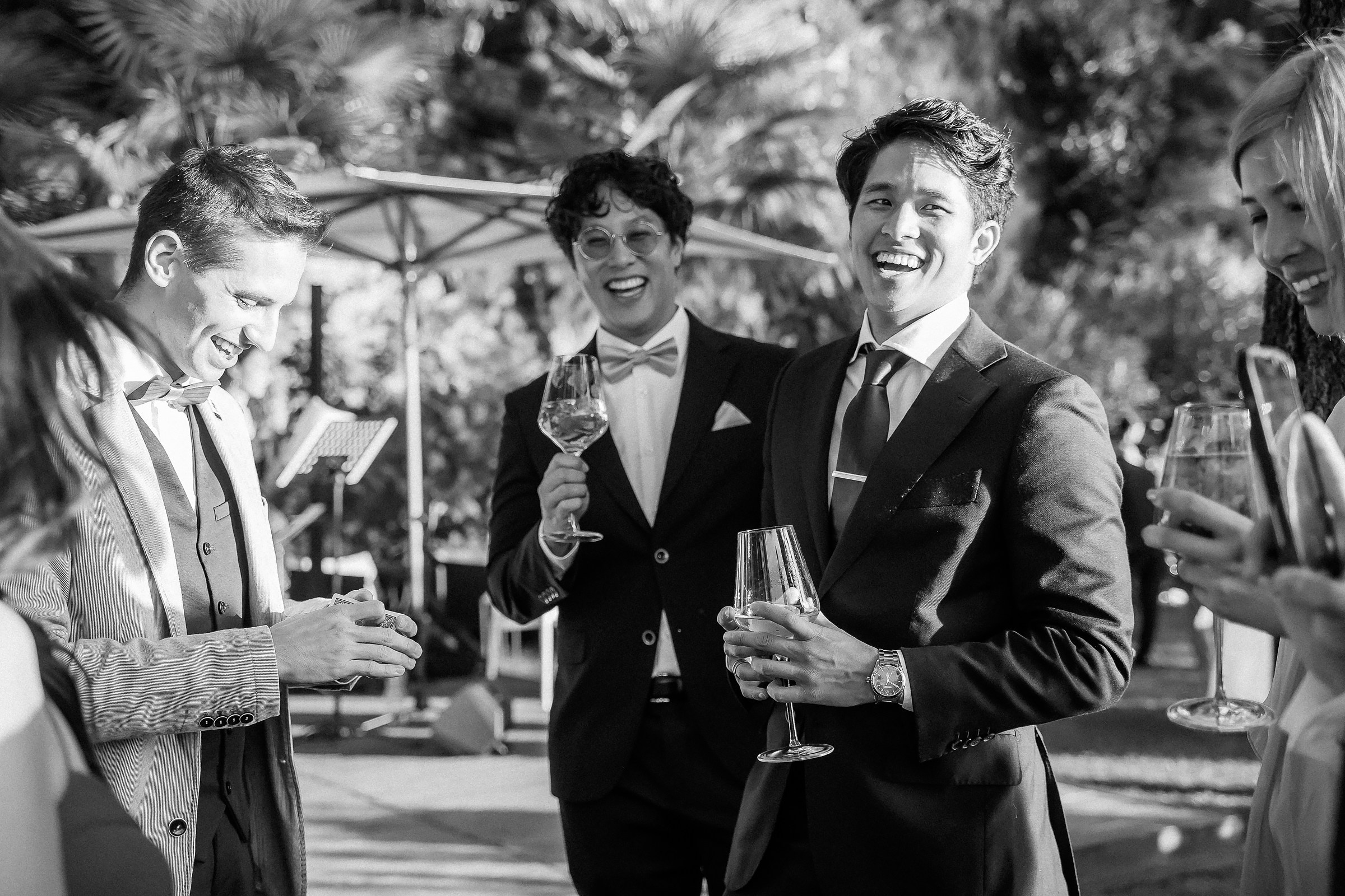 Three men and a woman in suits and dresses enjoy drinks, smiling and conversing amid a garden ambiance.