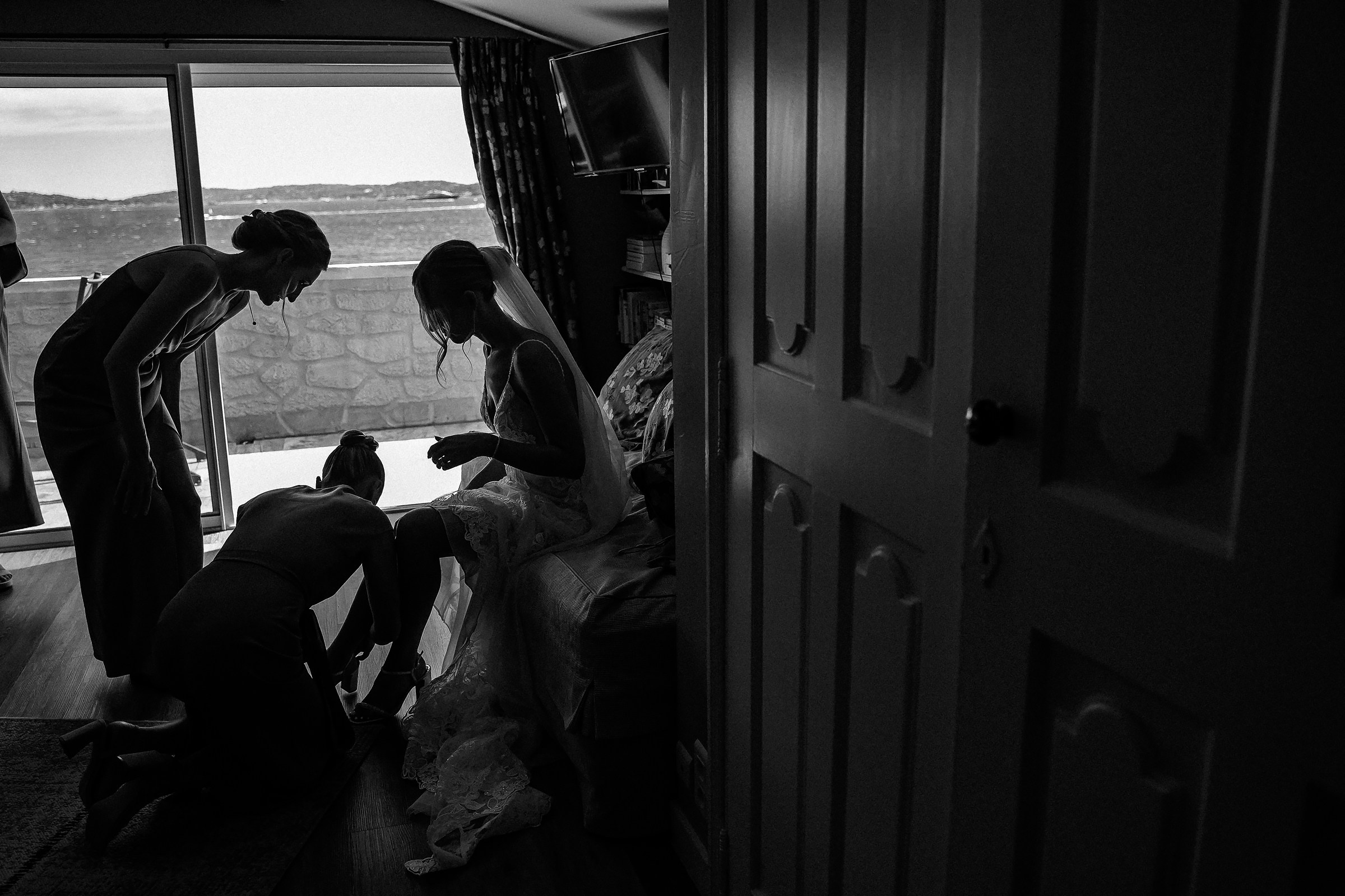 A silhouetted scene of the bride seated with two bridesmaids assisting, framed by a large window revealing a view of the sea.