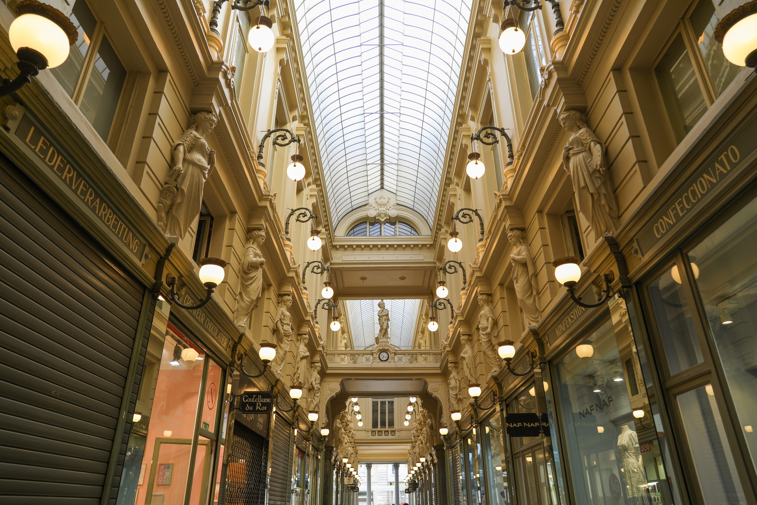 The interior view looking down the historic Passage du Nord in Brussels, a 19th-century covered shopping arcade with a glass ceiling, ornate yellow walls, statues, and shopfronts. Signage for "LEDERVERARBEITUNG" and "CONFECCIONATO" is visible.