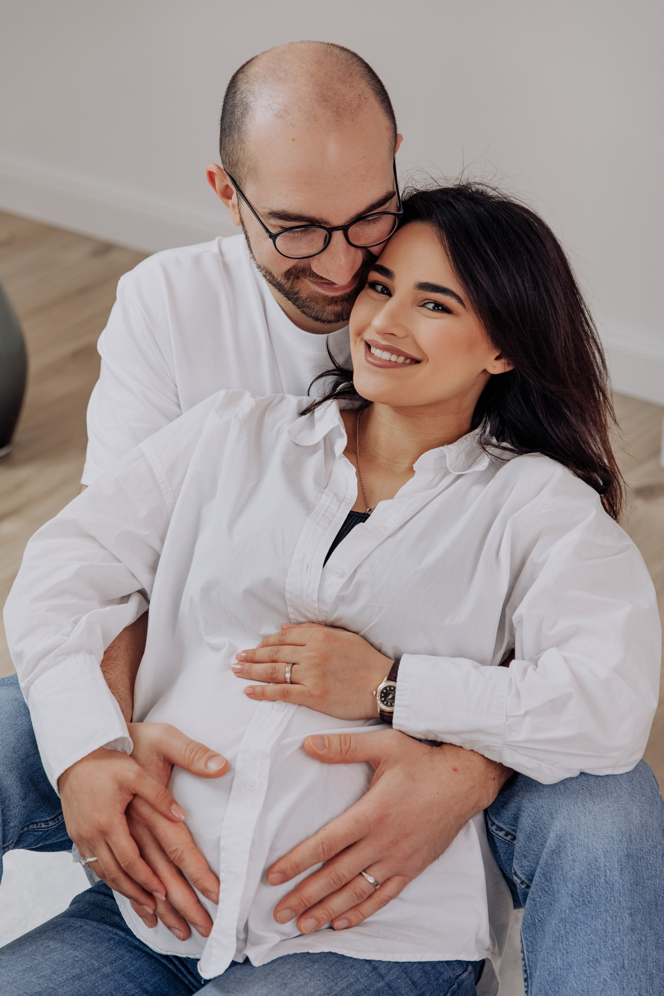 Mann umarmt seine schwangere Frau während eines Fotoshootings, beide lächeln glücklich.