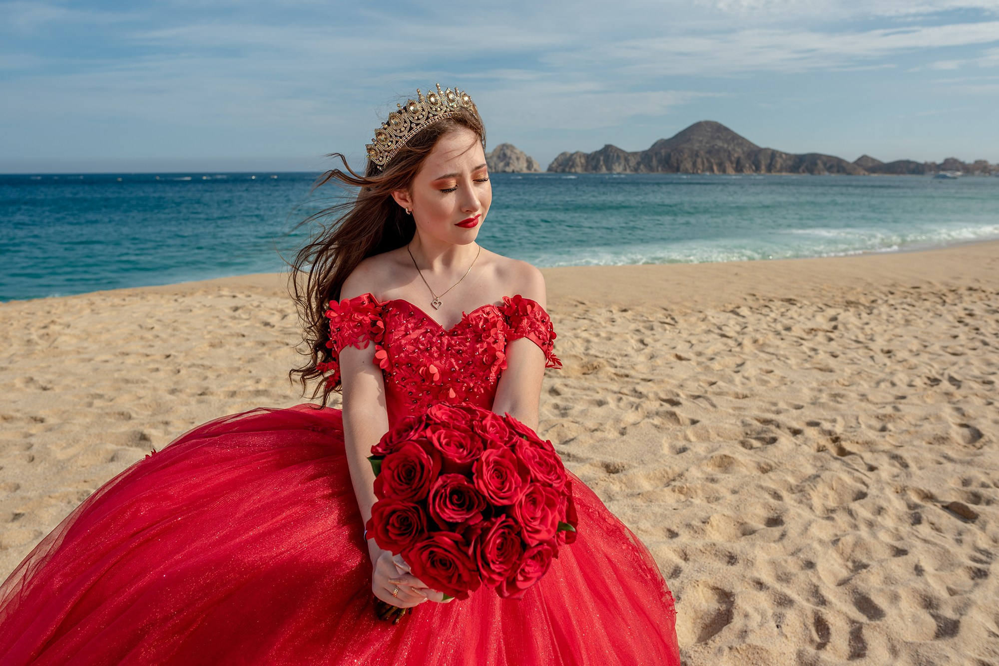 XV años photoshoot in Cabo San Lucas – Sofie wearing a stunning red quinceañera dress with a bouquet of red roses, photographed at El Médano beach with the Finisterra rock formations in the background. Destination XV photographer in Los Cabos