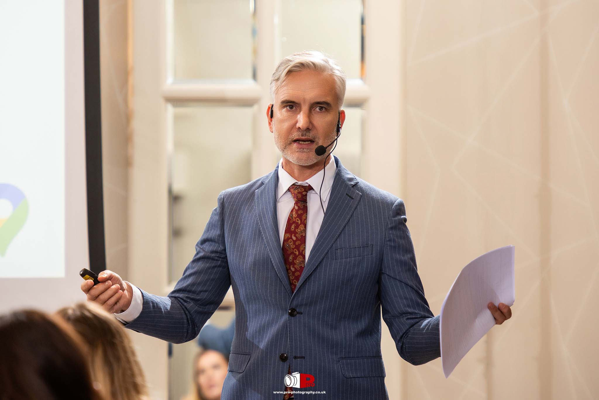 A confident male keynote speaker in a pinstripe suit presenting to an engaged audience at a corporate event.