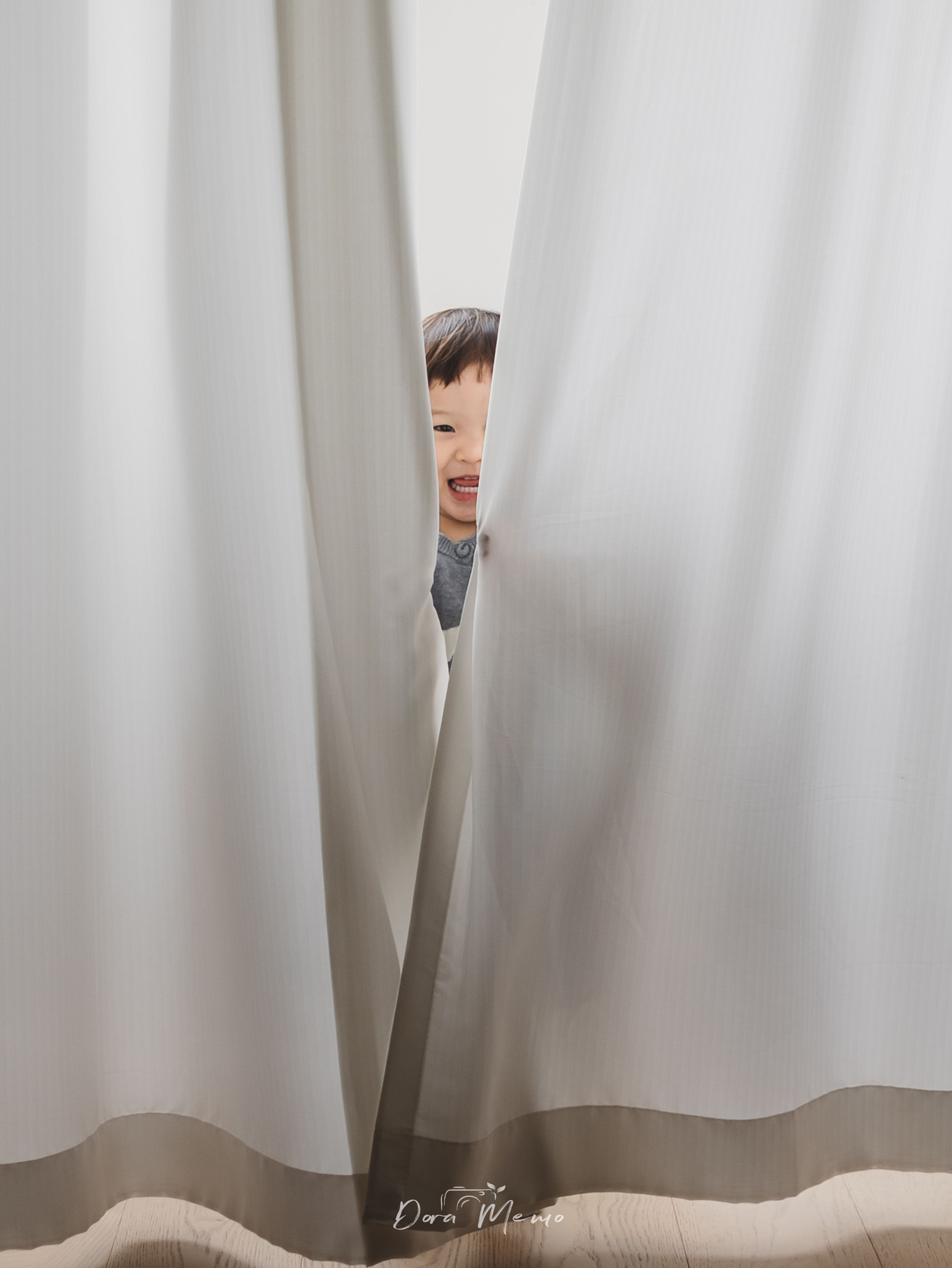 Playful toddler hiding behind sheer white curtains, peekaboo moment captured by a Shanghai family photographer at home.
