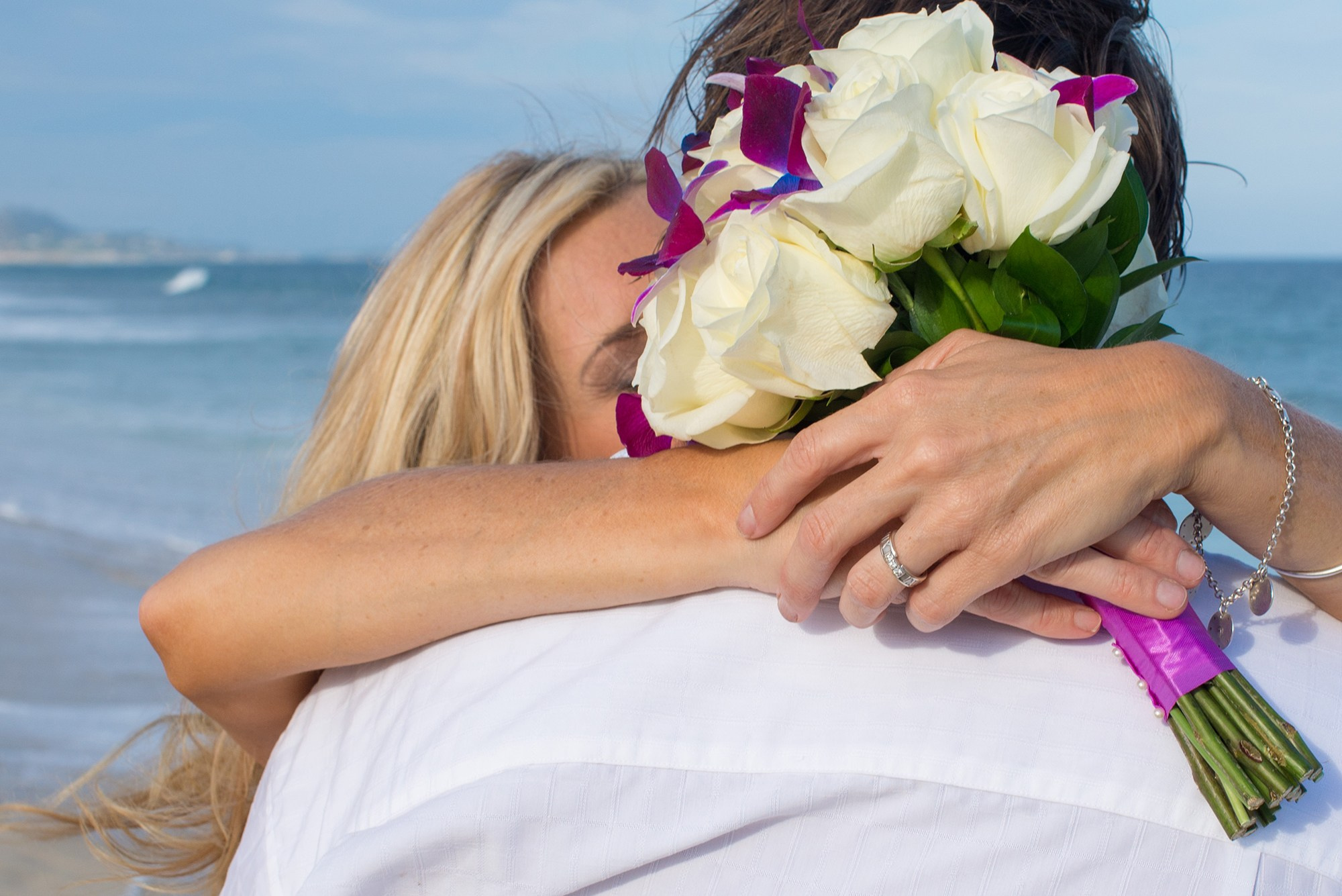 Beach wedding portrait in Los Cabos – bride embracing groom with bouquet and ring visible, cinematic sunset tones by the ocean