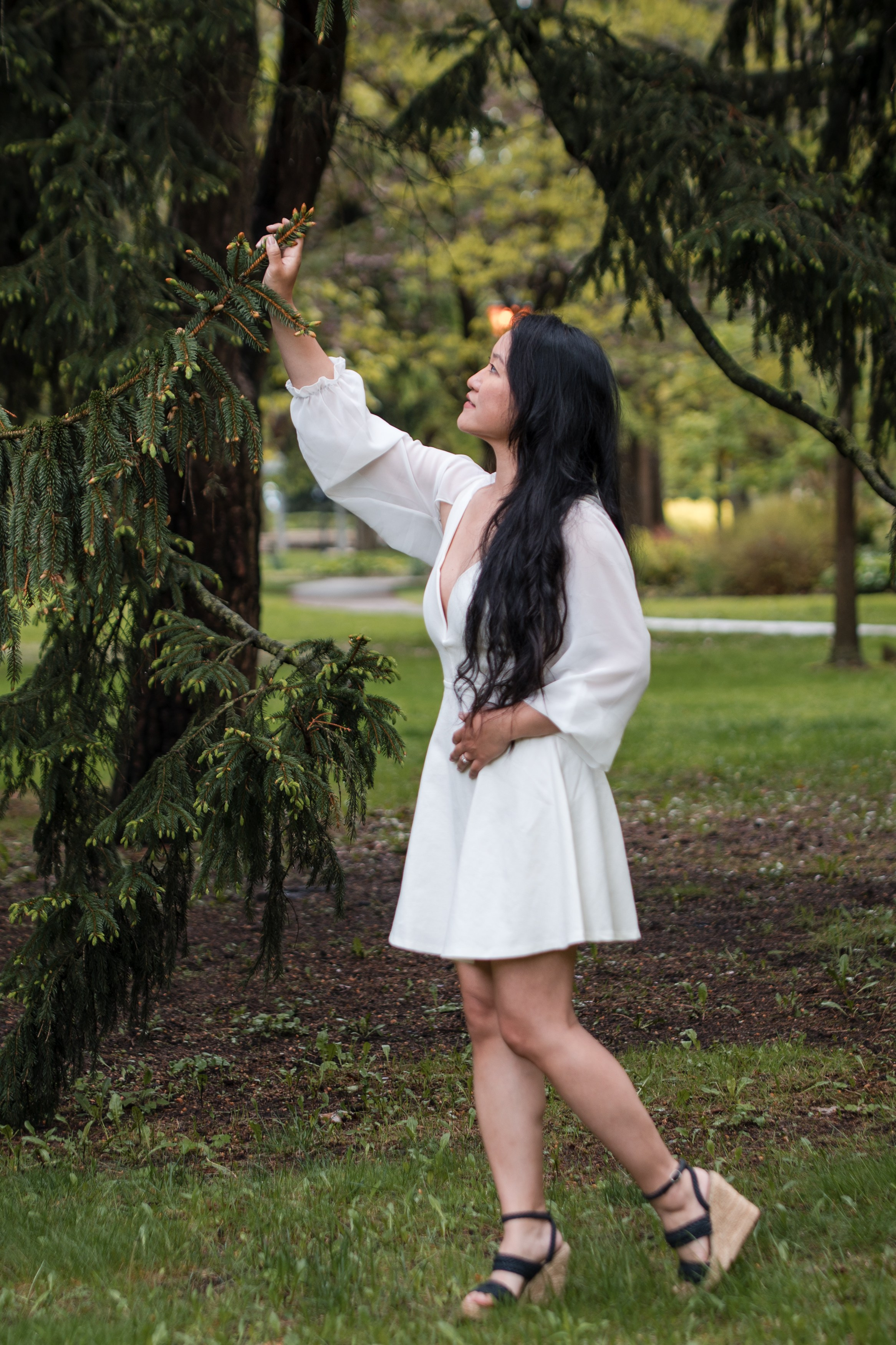 Bride-to-be in a white dress gently touching a pine tree branch during a forest engagement photoshoot.