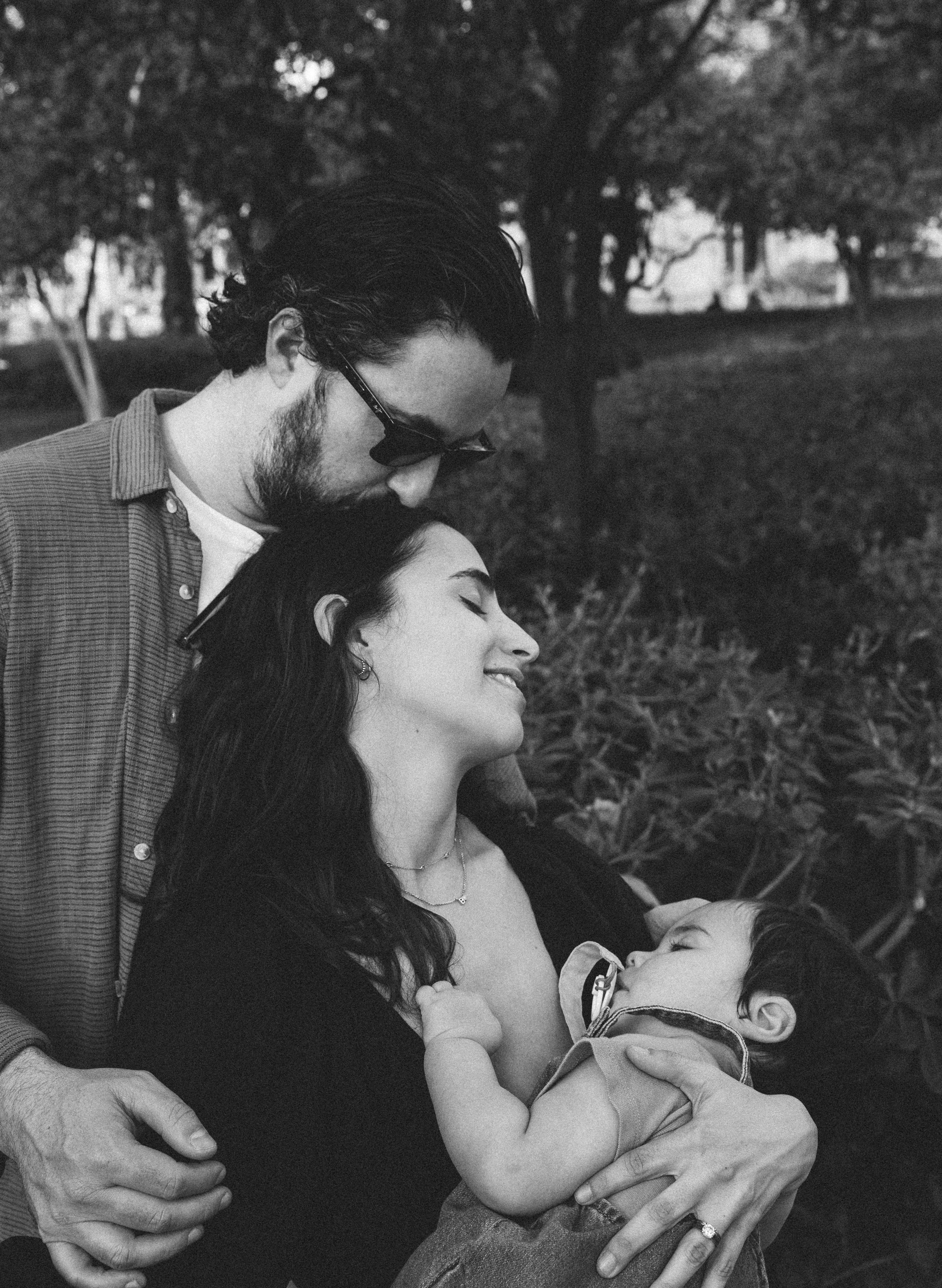 Retrato familiar en blanco y negro que captura un abrazo pacífico entre padres y su bebé dormido, rodeados de árboles en un parque tranquilo de Valencia, España — una escena serena llena de amor y conexión.