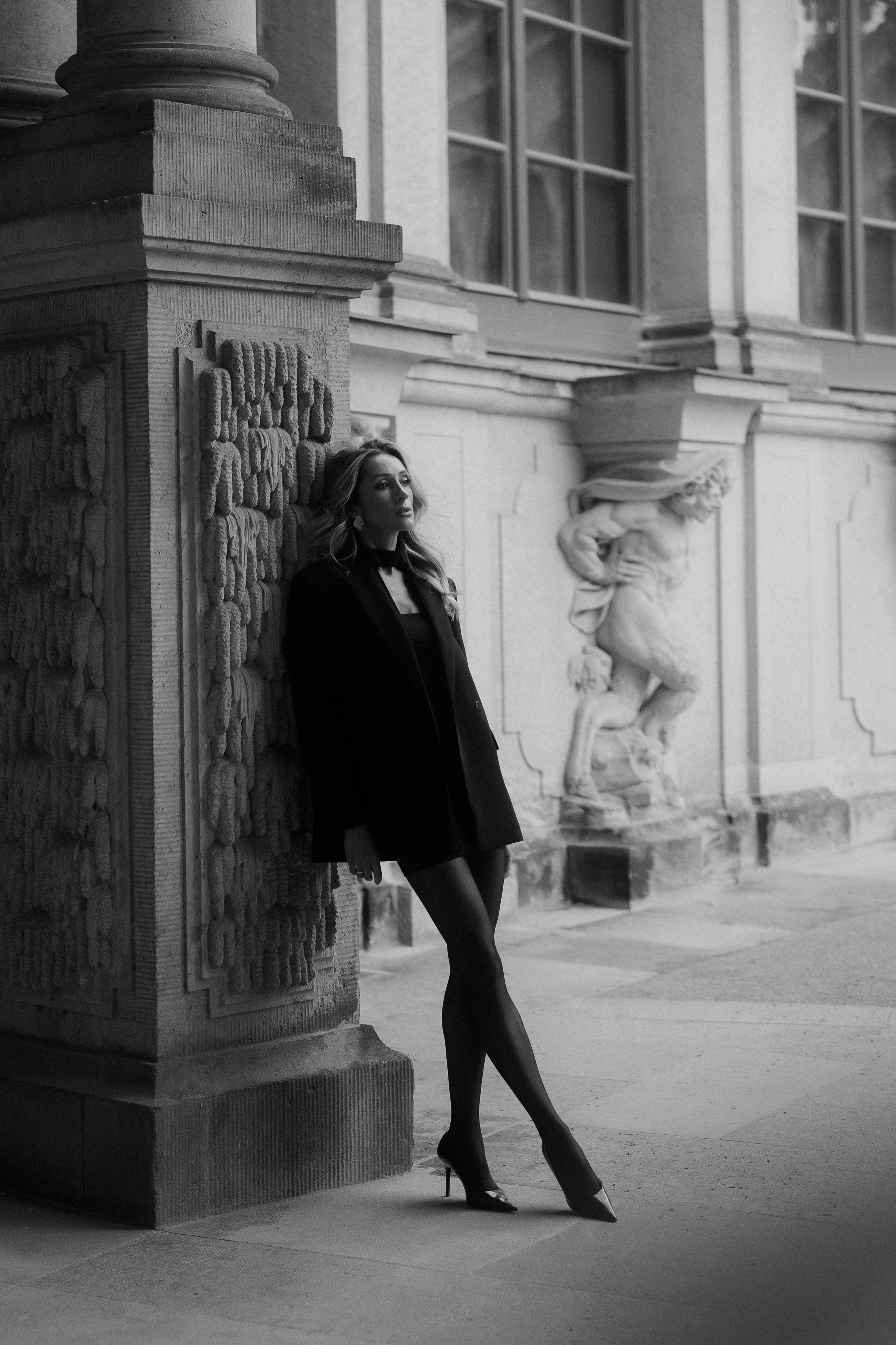 Black and white editorial portrait of an elegant woman in a black blazer and dress leaning against historic architecture in Dresden, Germany. Fine art female portrait photography by a photographer based in Leipzig and Wurzen. Чёрно-белый редакционный портрет элегантной девушки в чёрном жакете и платье у исторической колонны в Дрездене, Германия. Художественная женская портретная фотография от фотографа из Лейпцига и Вурцена.
