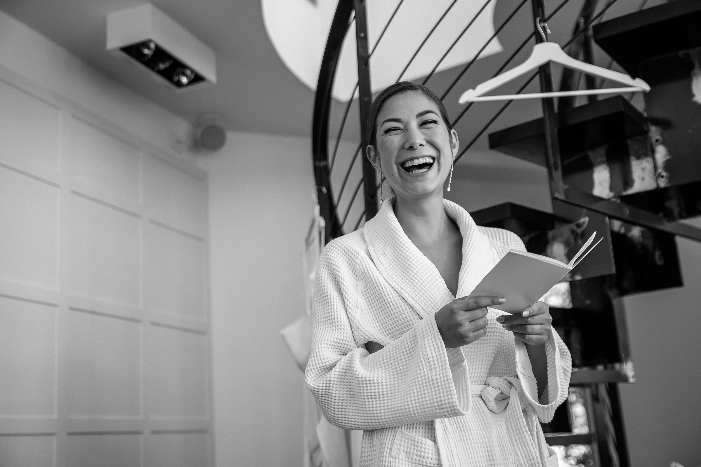 Bride in a robe laughs while reading a note near a spiral staircase.
