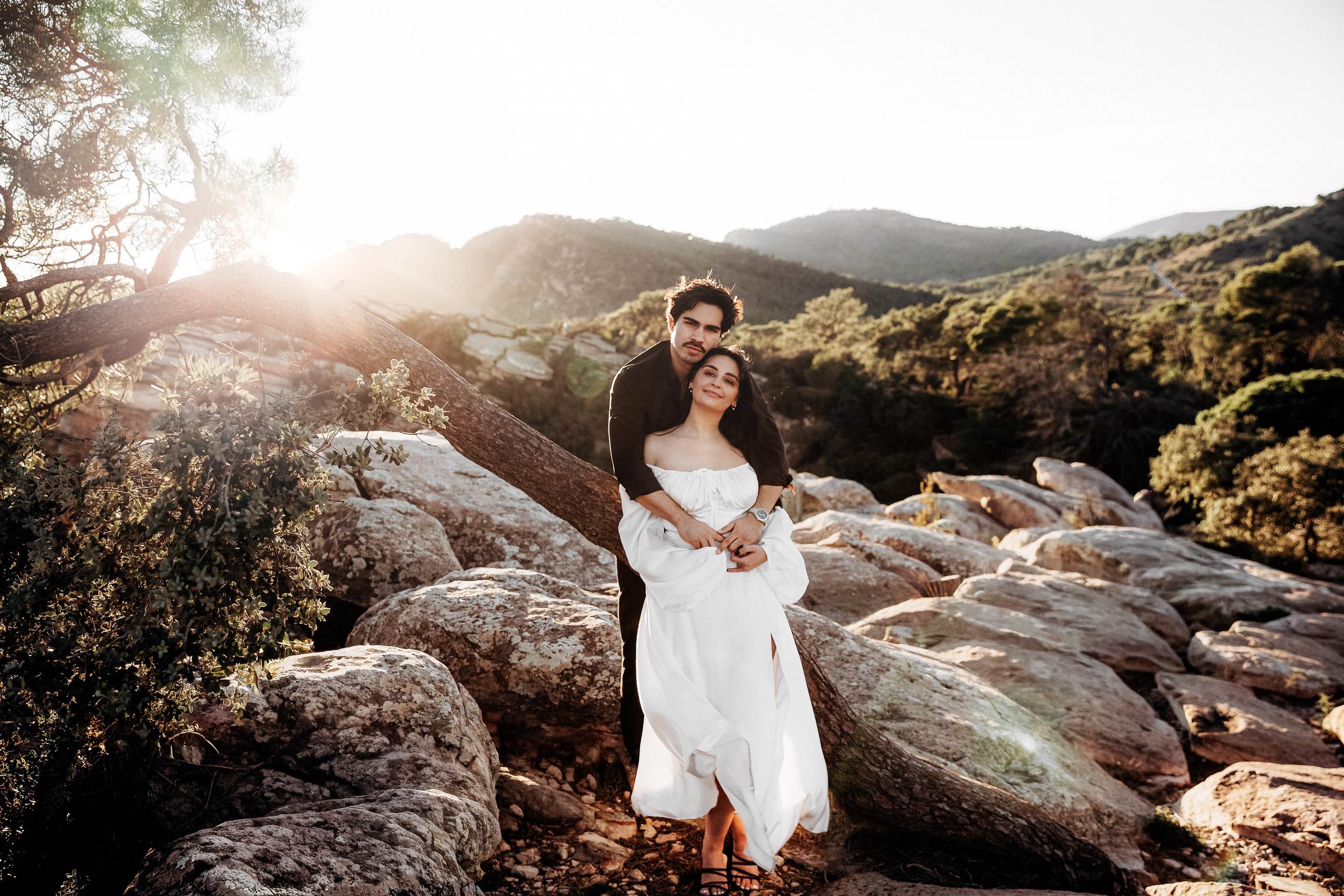 Romantic engagement photoshoot in València, Spain, showing a couple embracing on sunlit rocky hills surrounded by Mediterranean nature, with soft golden-hour light and mountains in the background, capturing an intimate and cinematic pre-wedding love story — perfect for engagement photography, wedding storytelling, elopements, and professional engagement and wedding photoshoots in València and across Spain.