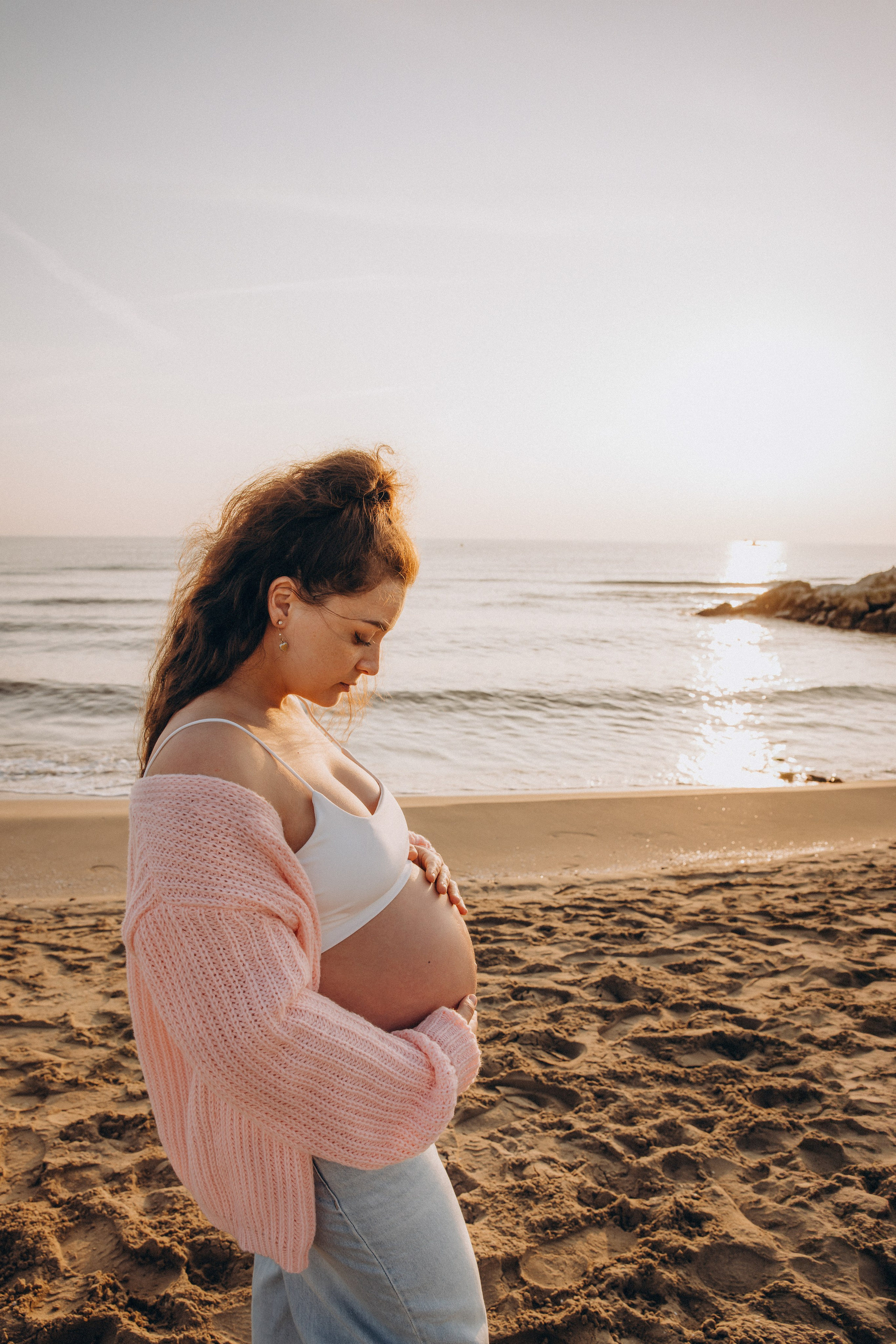 Sesión de fotos de embarazo en Valencia, España, con una mujer embarazada caminando por la playa con la suave luz del atardecer, creando un momento tranquilo y cinematográfico — ideal para sesiones de embarazo y maternity en Valencia y en toda España.