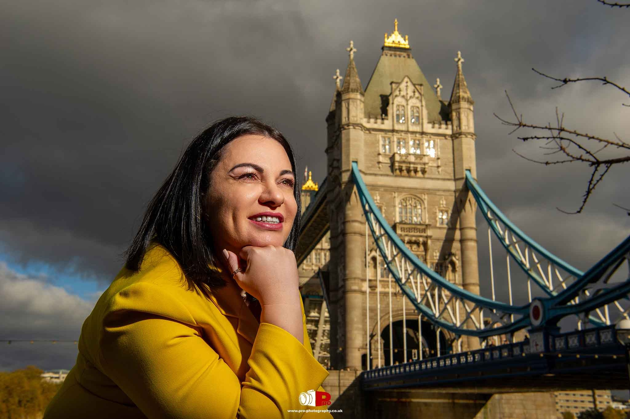 A smiling woman in a yellow blazer posing with London's iconic Tower Bridge in the background.