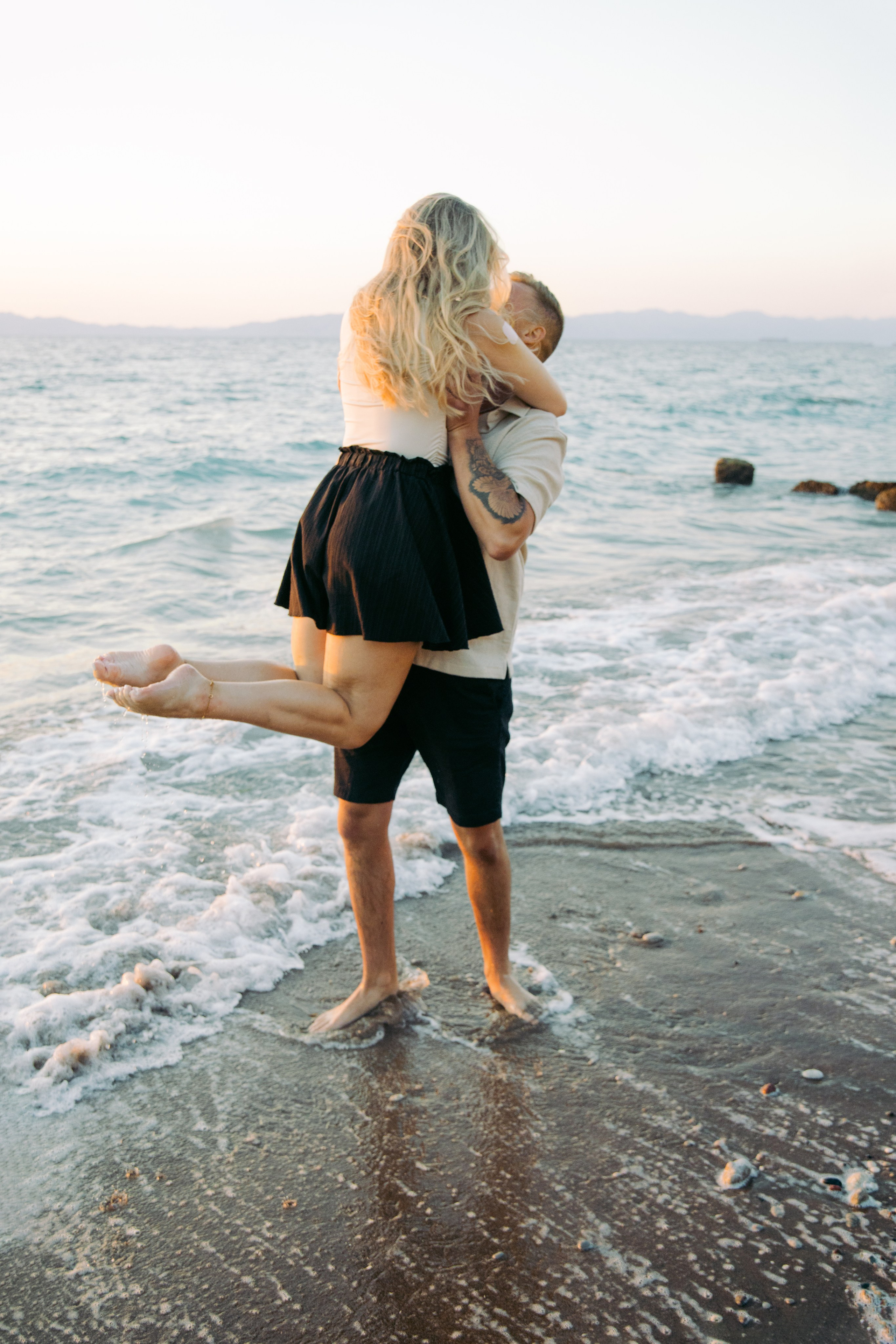 "Ein Paar spaziert Hand in Hand am steinigen Ufer von Kato Petres auf Rhodos entlang. Sie gehen direkt am Wasser, während im Hintergrund die hohen, zerklüfteten Kalksteinfelsen aufragen und das blaue Meer sanft gegen die Kiesel brandet."