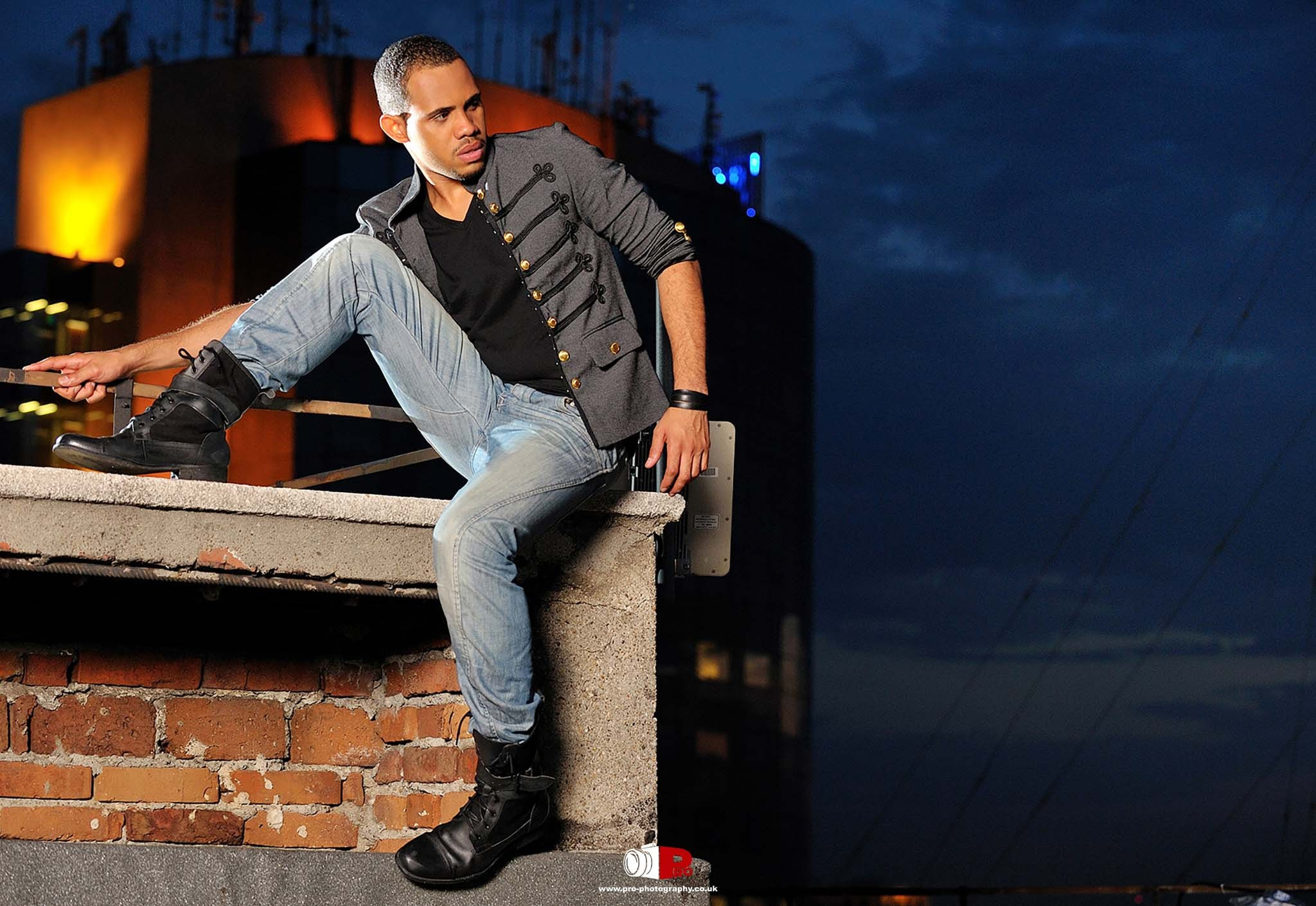 A young man in casual fashion posing on a rooftop with a city skyline illuminated behind him at dusk.