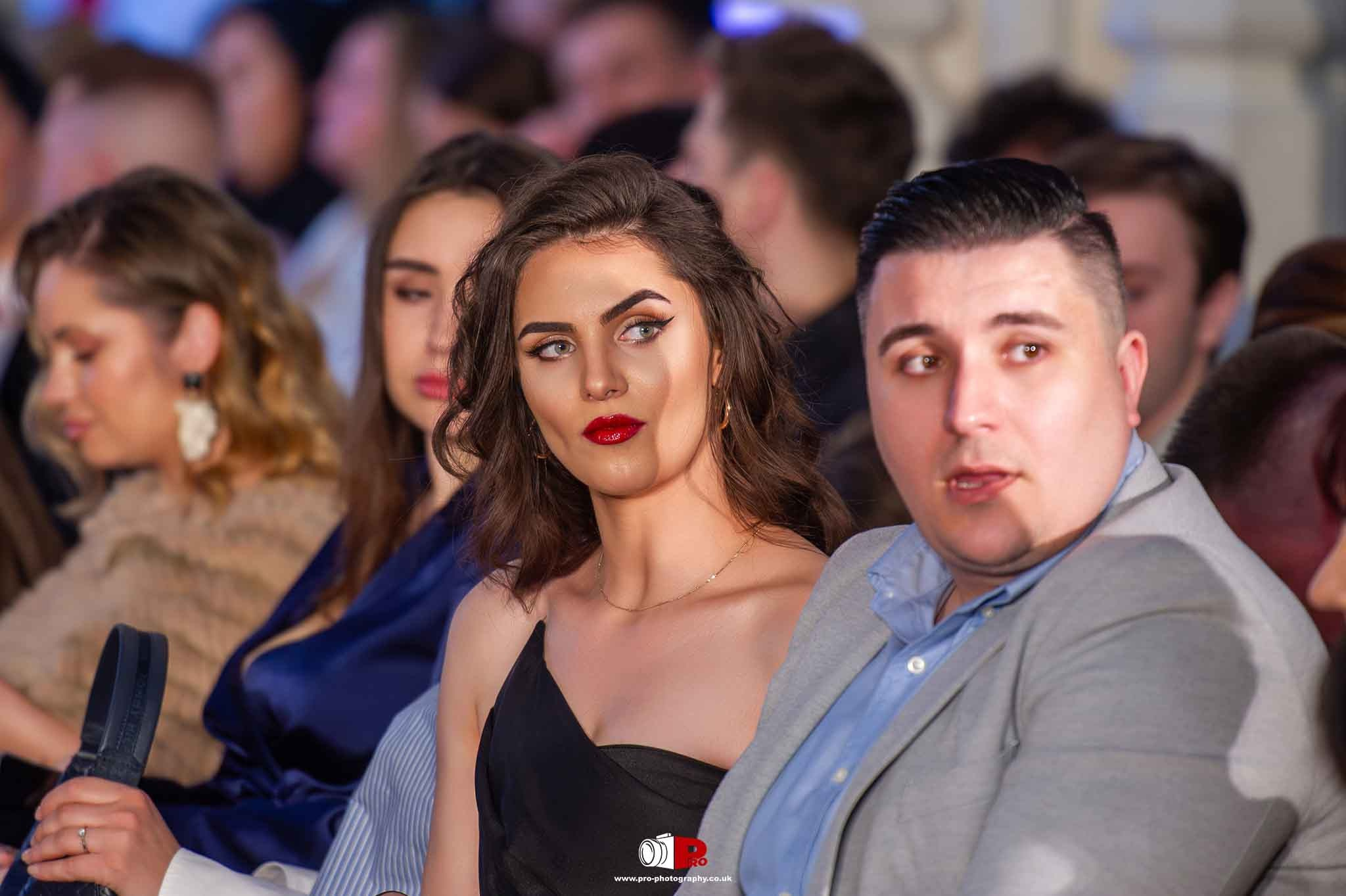 An elegant woman in a black dress and red lipstick conversed with a man in a grey suit at a gala event.