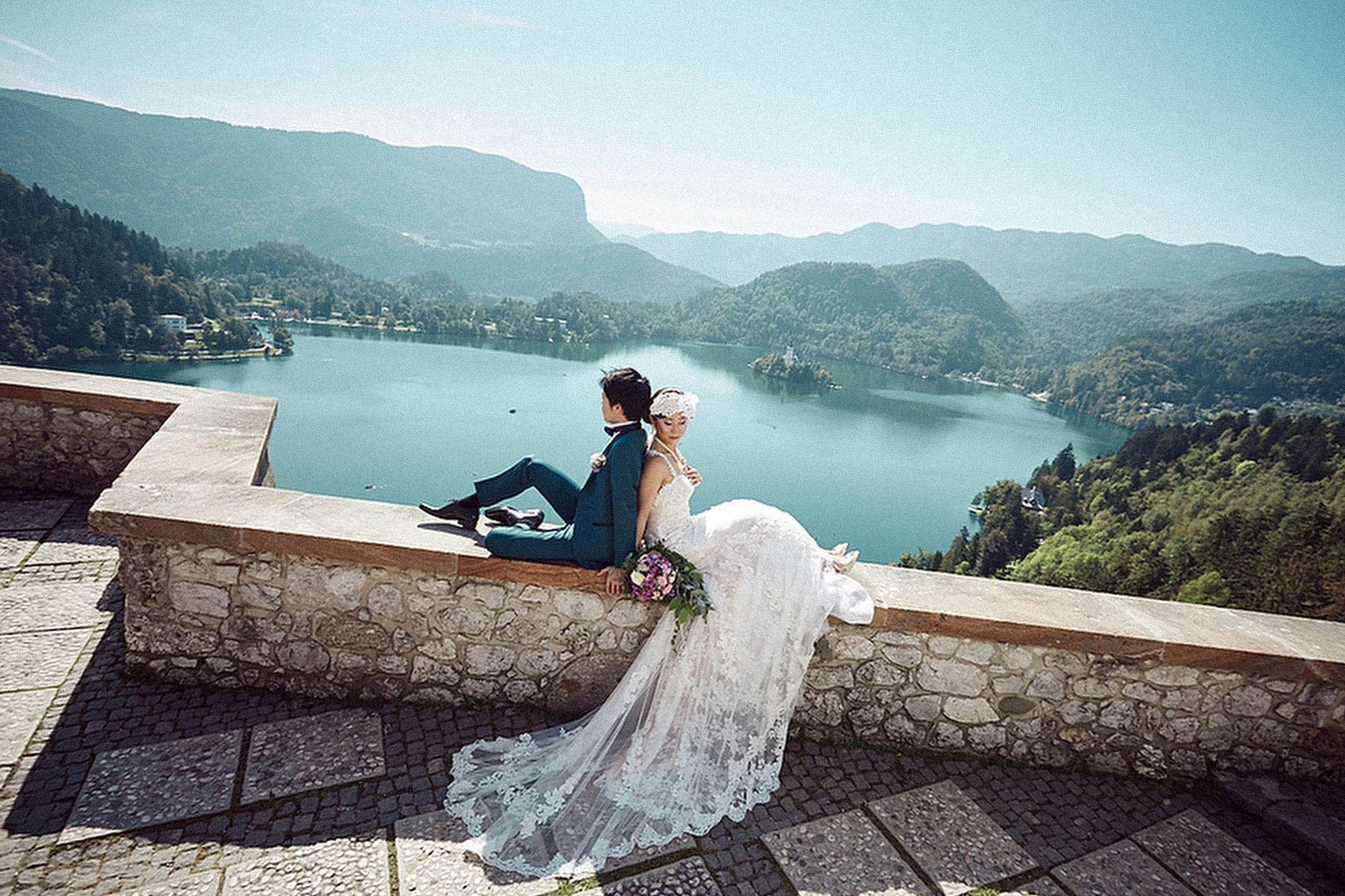 Newlyweds portrait sitting on medieval wall Bled Castle overlook.