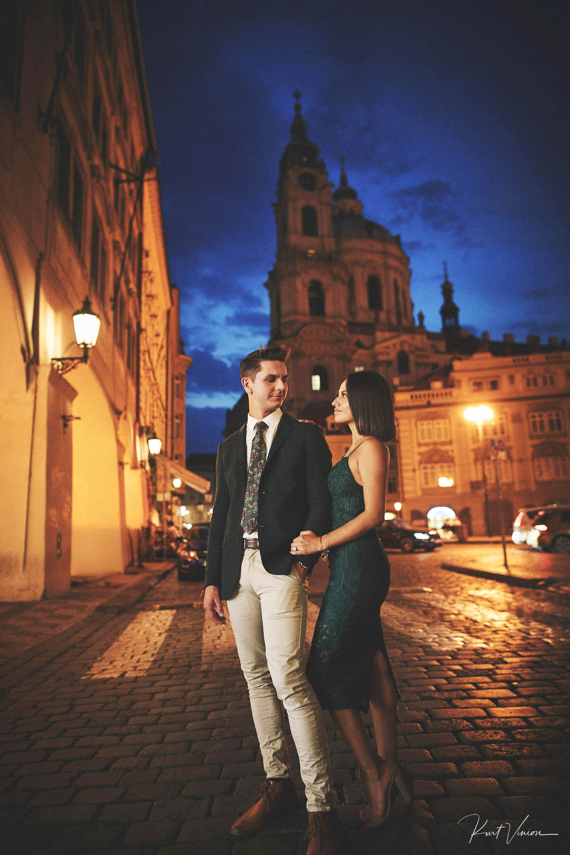 Woman in green dress holding partner’s arm in Mala Strana at night after engagement.