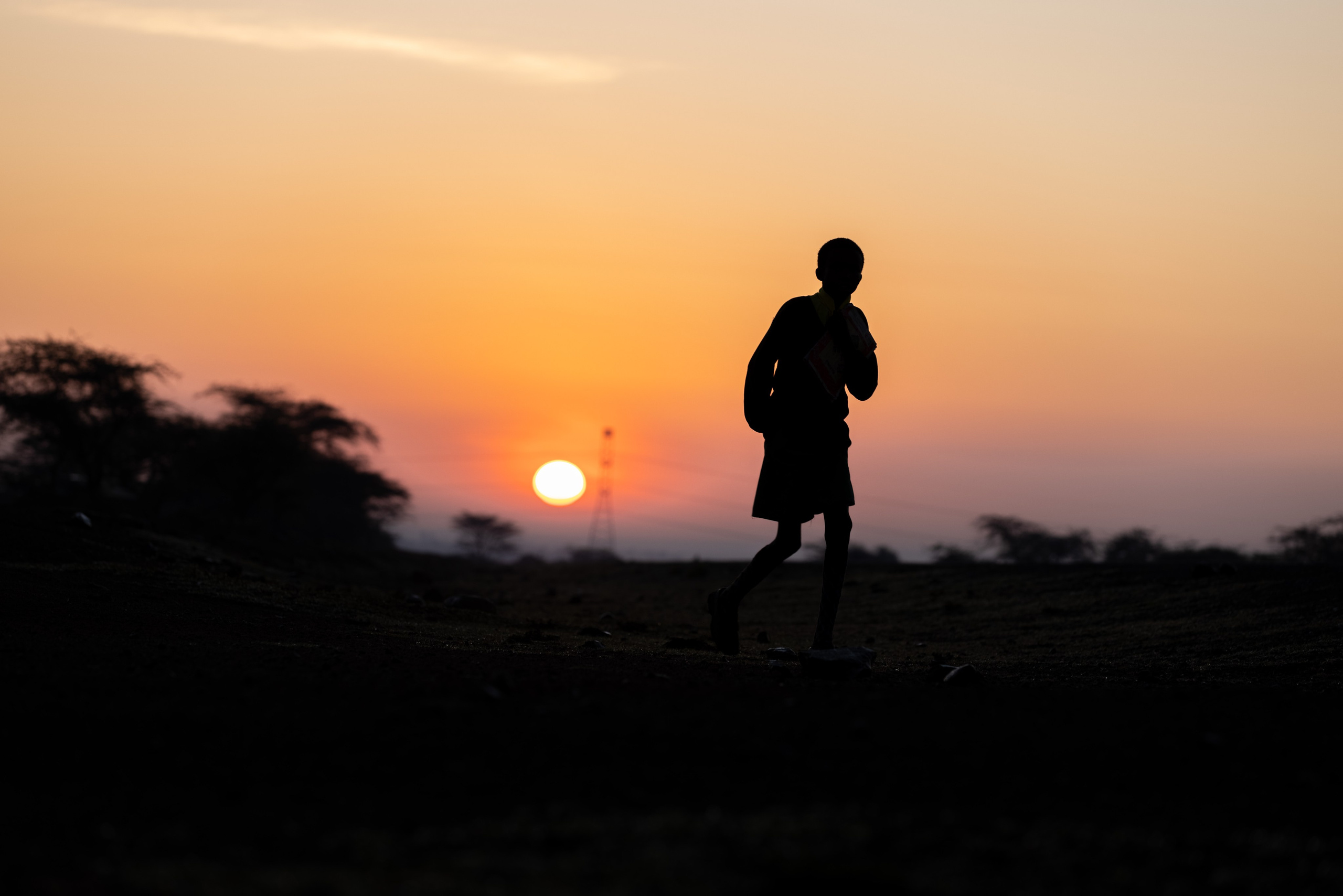 A Silhouette full shot of a child going to school. In the background is the sunrise peaking in the horizon. Documentary photography 