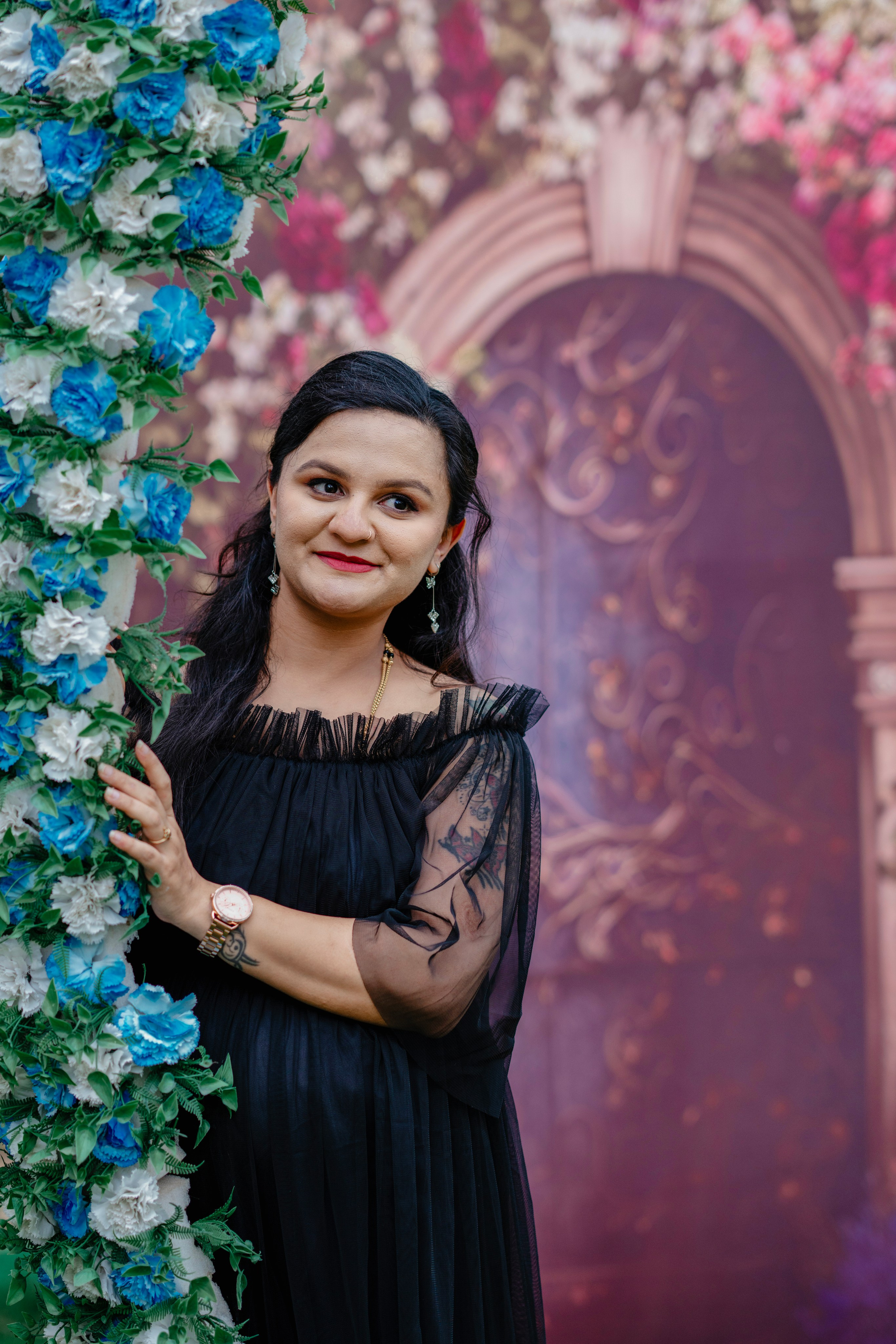 Maternity photoshoot in Bengaluru studio featuring a woman in a black off-the-shoulder tulle gown posing against a floral garden arch backdrop.