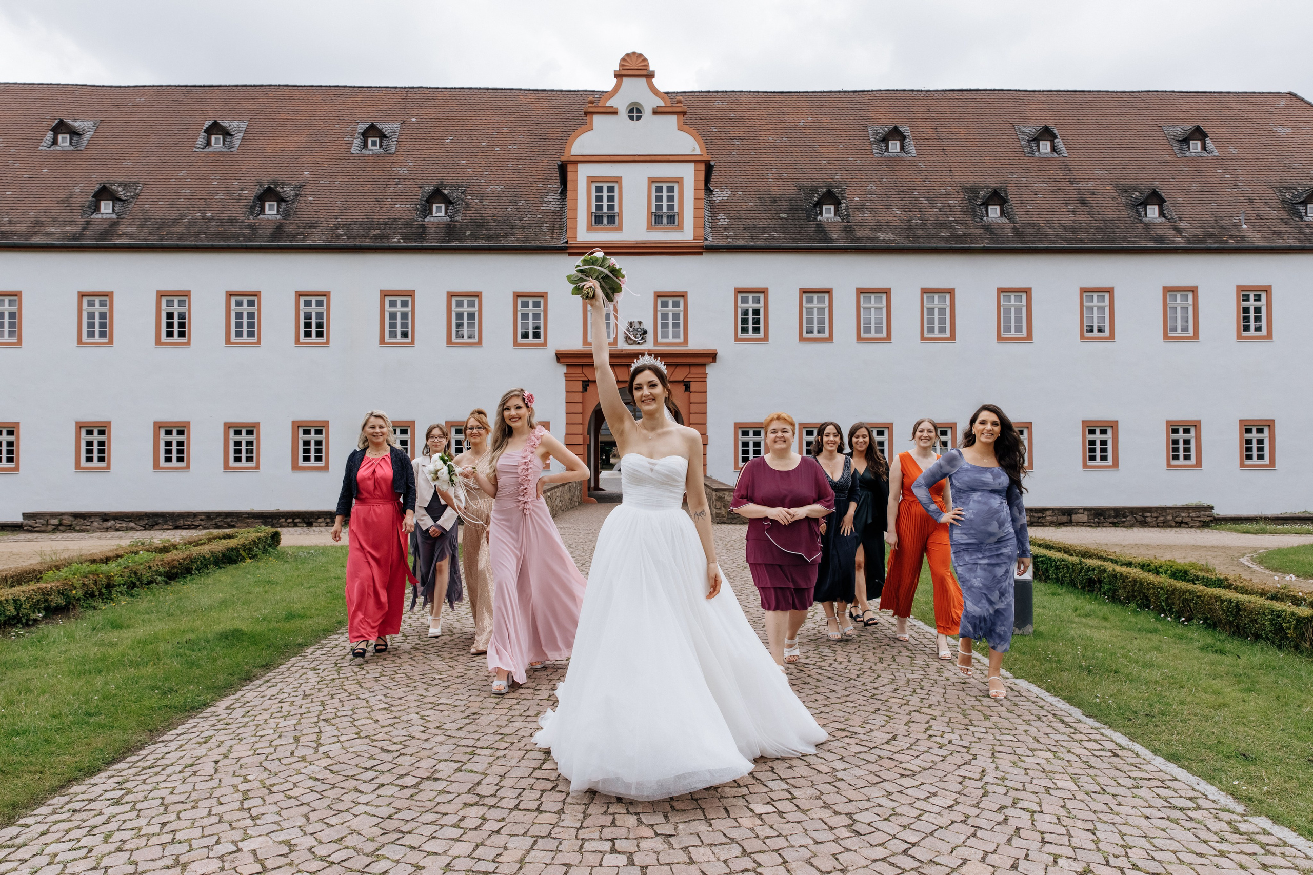 Die Braut hält ihren Brautstrauß nach oben und geht nach vorne, während ihre Freundinnen ihr folgen.