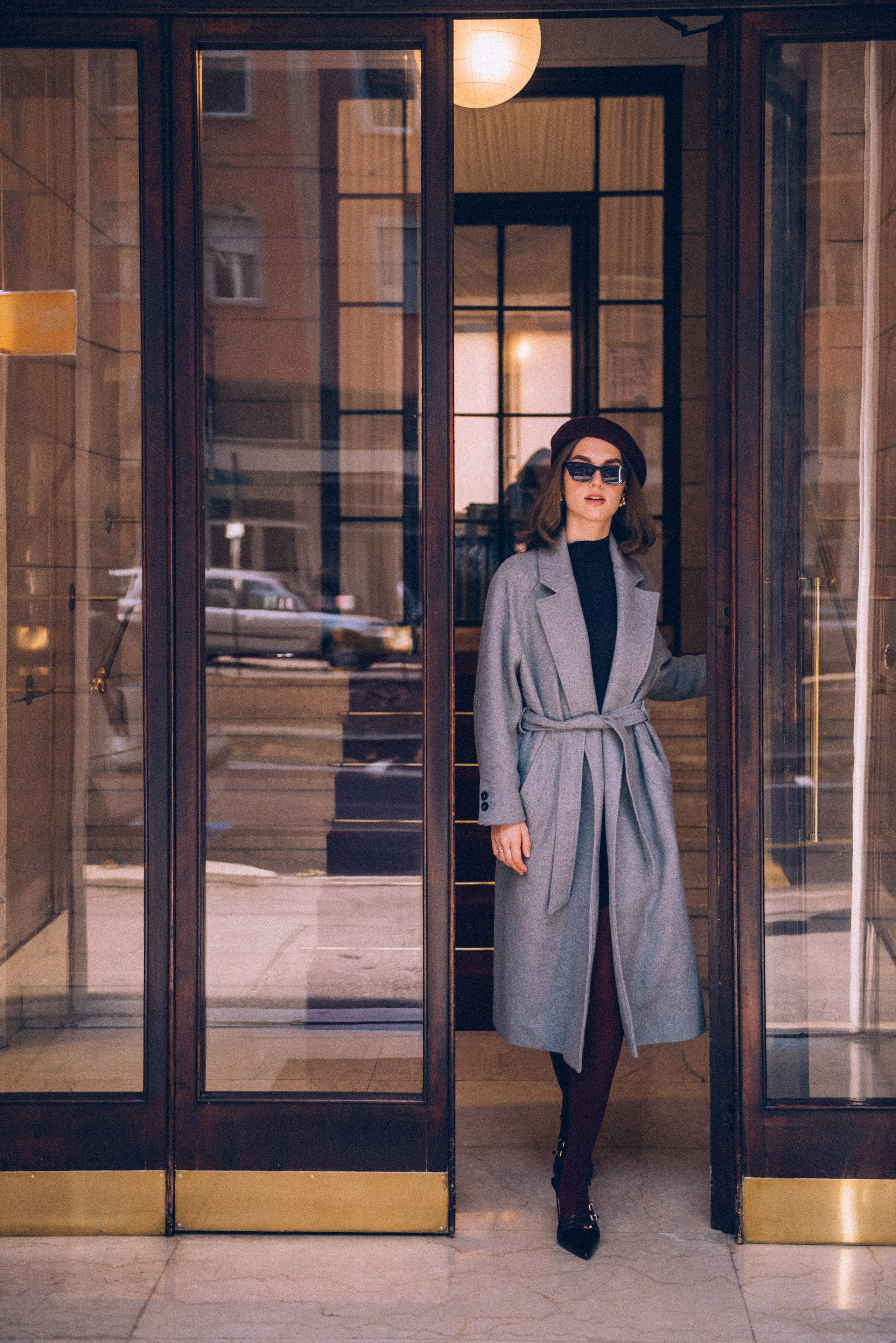 Chic Milanese street style: a stylish woman in a gray coat and burgundy tights stepping through a classic glass doorway, reflecting urban sophistication