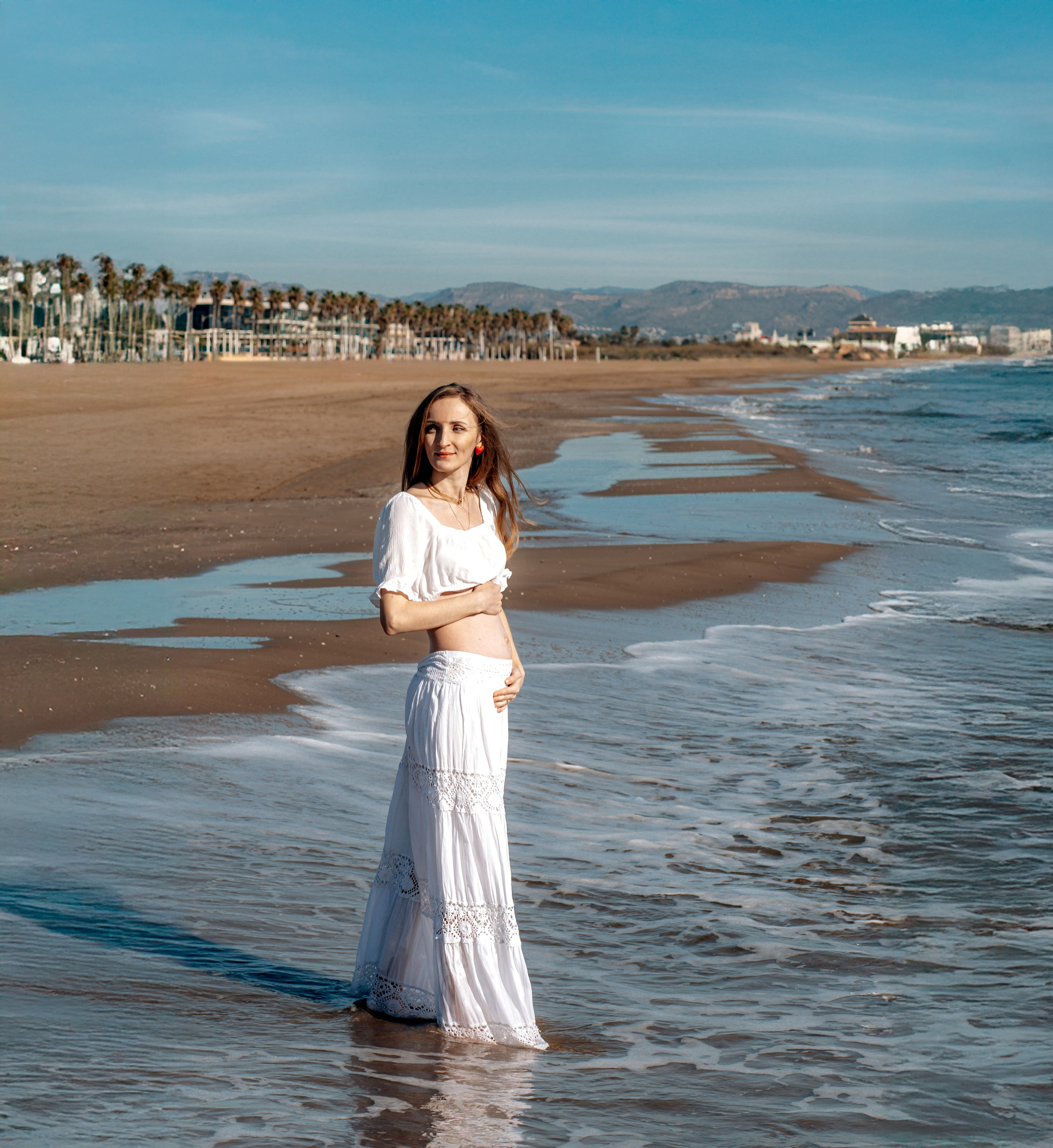Sesión de fotos de embarazo en València, España, con una mujer embarazada de pie y descalza en las suaves olas de una playa mediterránea de arena, sosteniendo delicadamente su barriga, capturada con luz natural suave, una atmósfera costera serena, palmeras en la orilla y montañas a lo lejos — ideal para fotografía de embarazo, sesiones de maternity, sesiones de embarazo en la playa, retratos artísticos de embarazo y sesiones naturales de embarazo en València y en toda España.