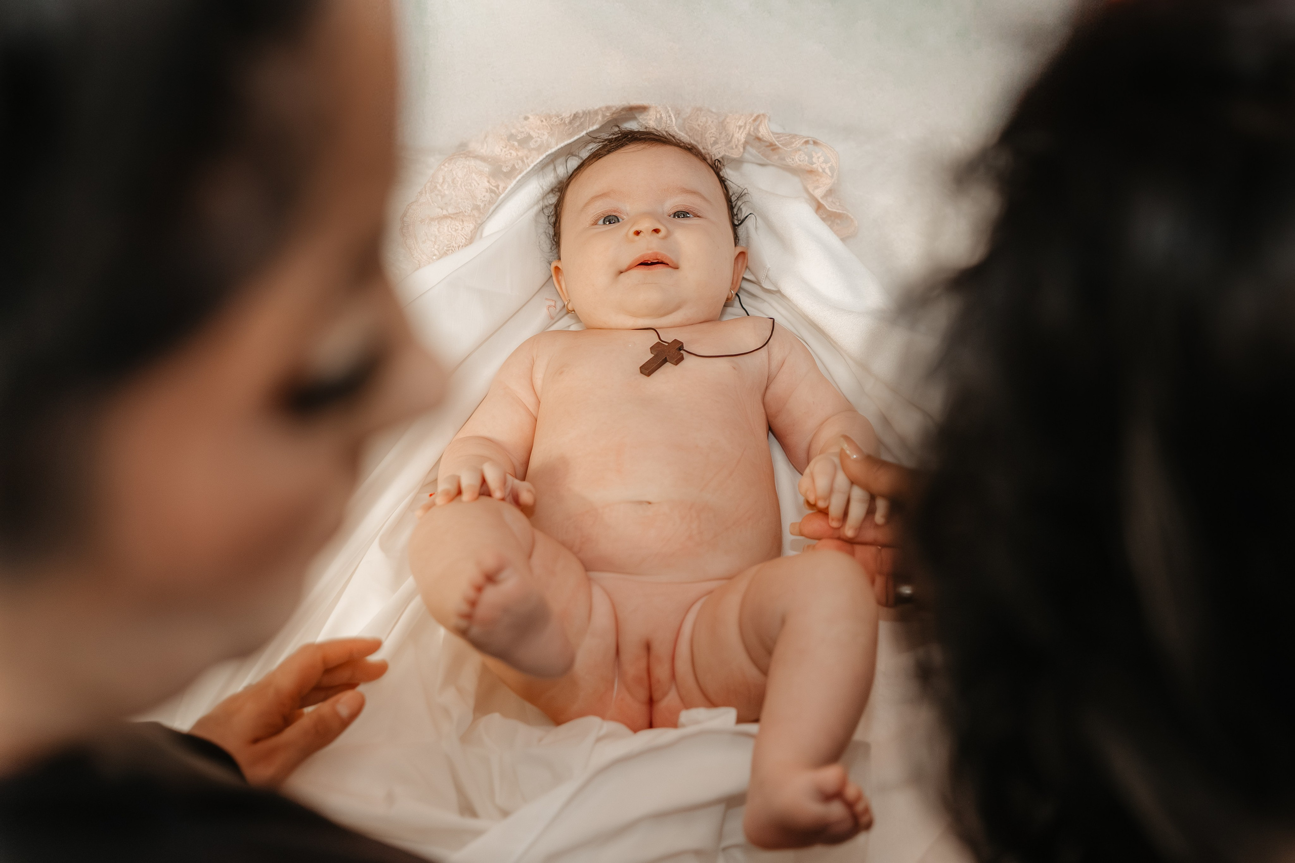 Înainte de a fi îmbrăcată în hainele noi, pruncul Eveline Maria stă pe masa de botez. Nașa și moașa (nasa) sunt încadrate în plan apropiat, pregătind momentul. Fotografie intimă a tranziției de la ritual la viața obișnuită.