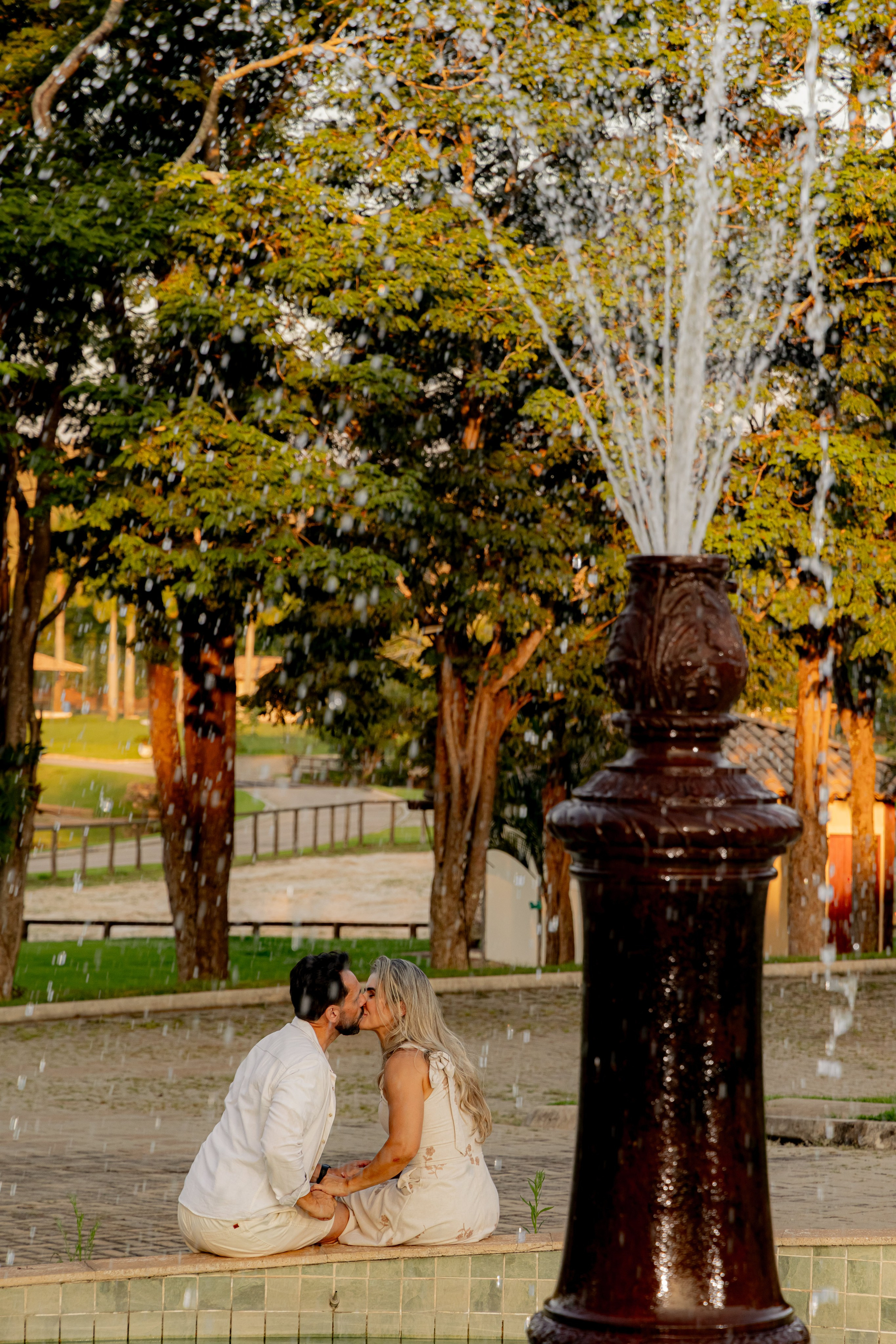 Casal sentado conversando ao lado do chafariz do Resort das Oliveiras, com gramado e lago ao fundo no final da tarde