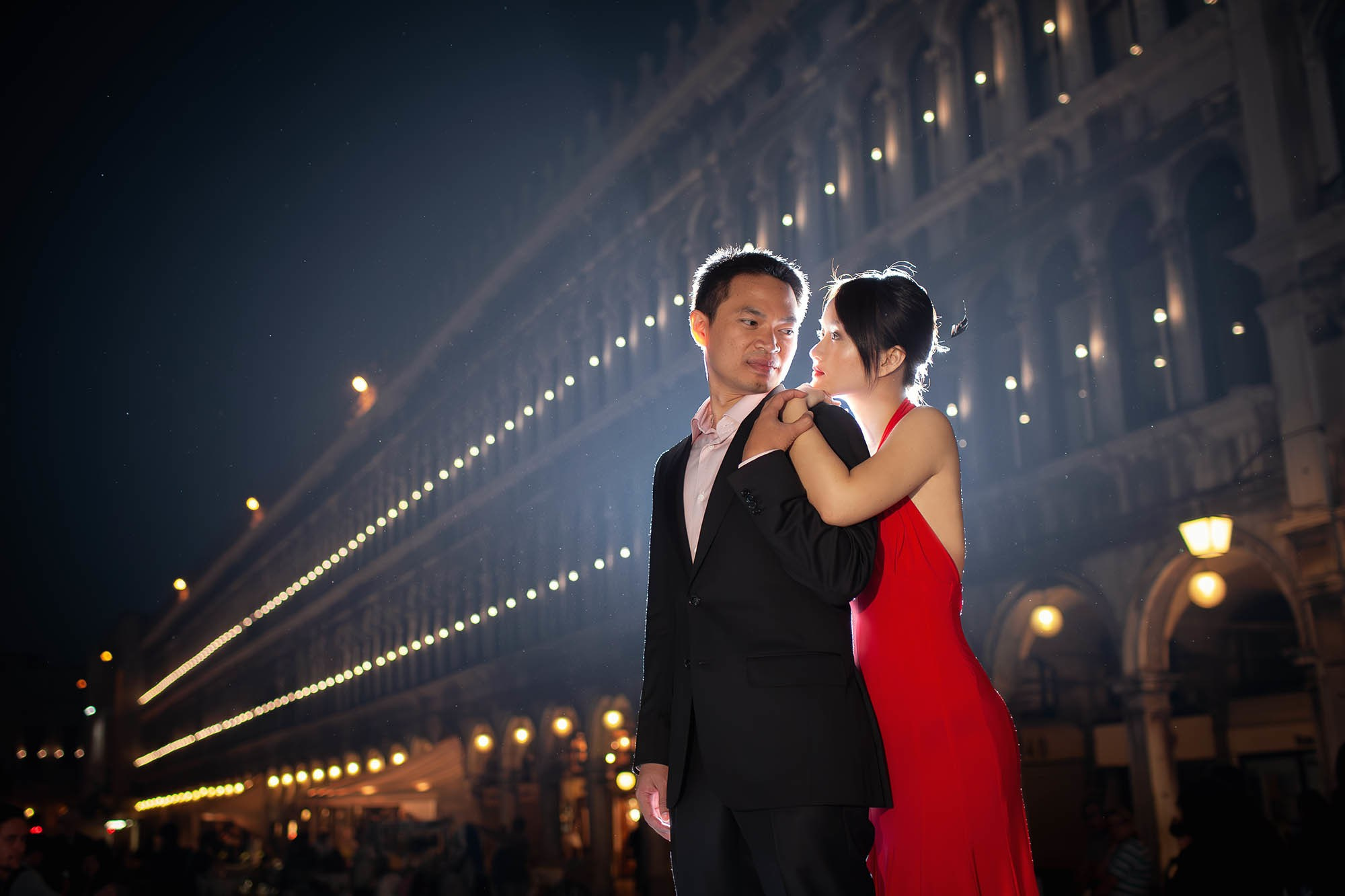 Woman in red dress embracing partner from behind in San Marco square Venice at night.