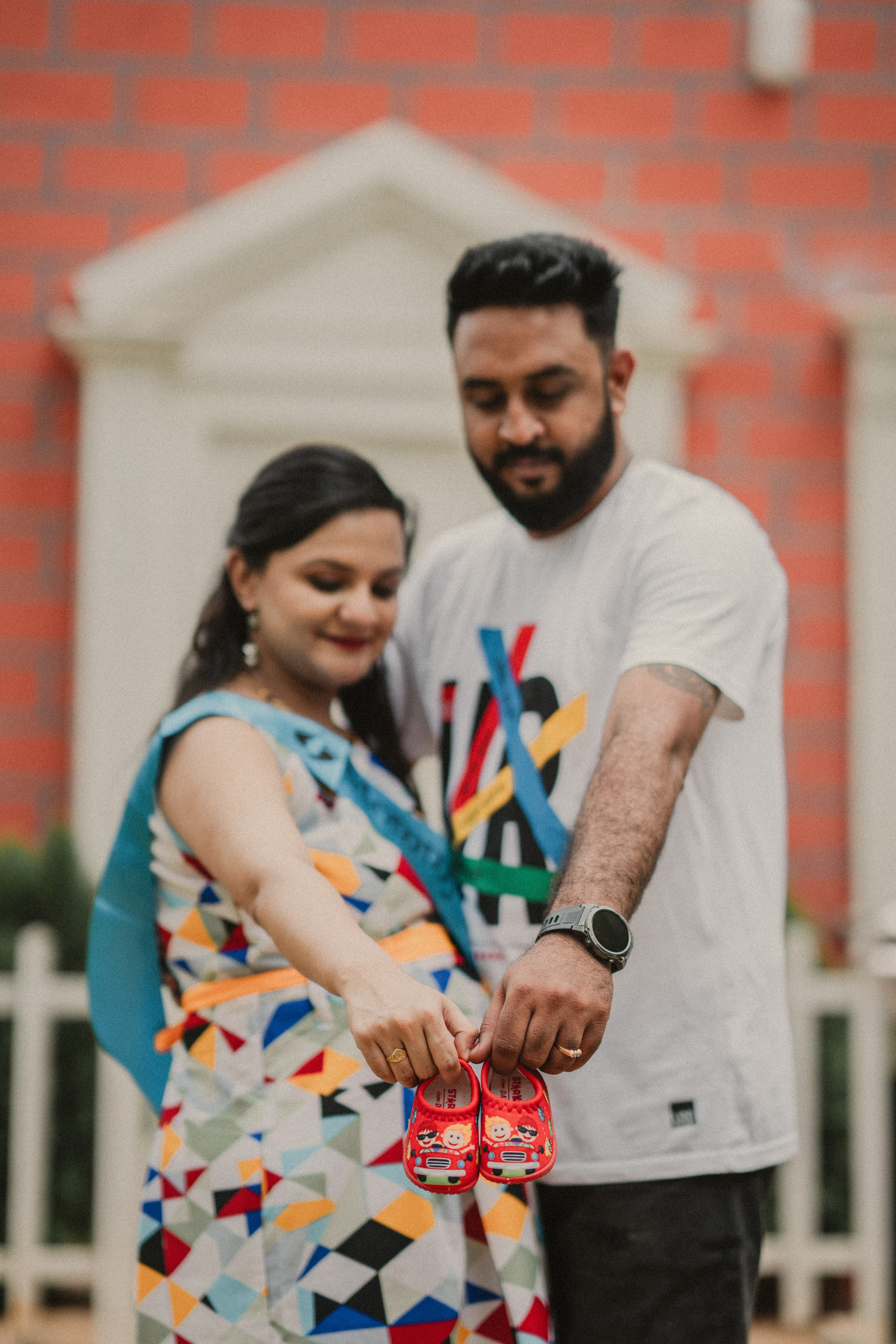 Maternity photoshoot in Bengaluru featuring a man and woman holding tiny red baby shoes in front of a colorful brick wall and white fence.