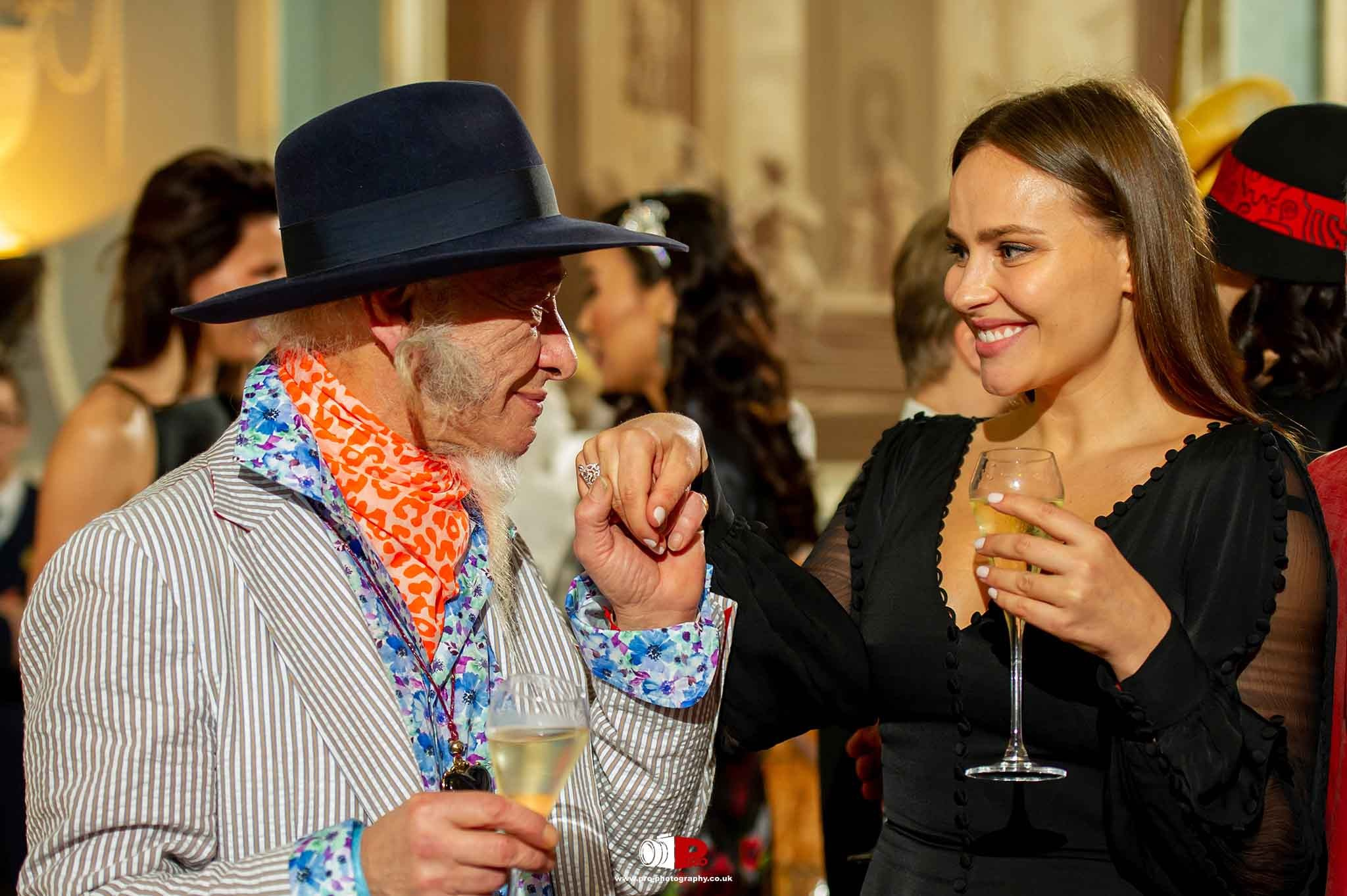 An elderly man in a stylish striped suit and colorful accessories shares a toast with a young woman in a black dress at a social event.