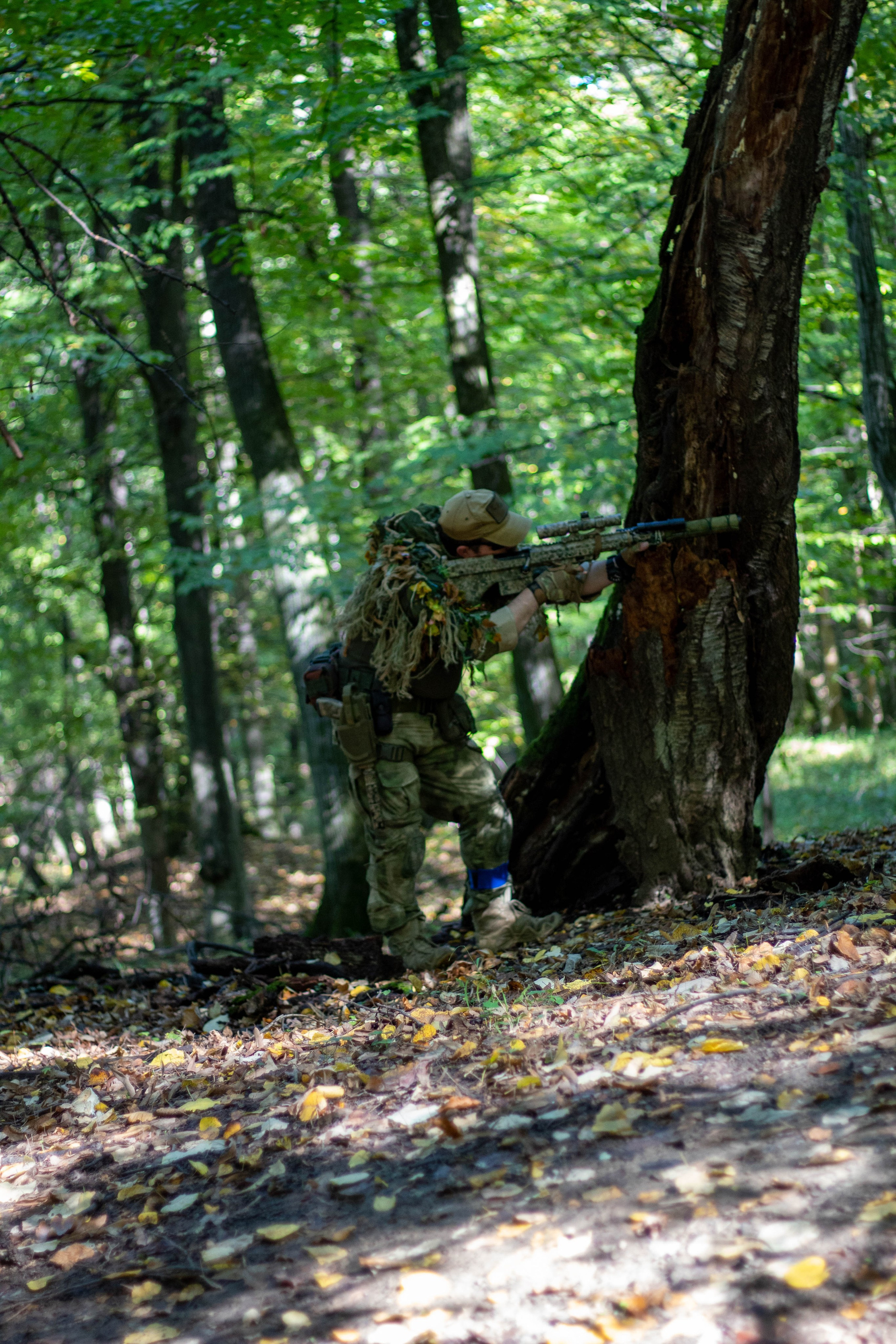 Player wearing full camouflage gear standing between tall forest trees.