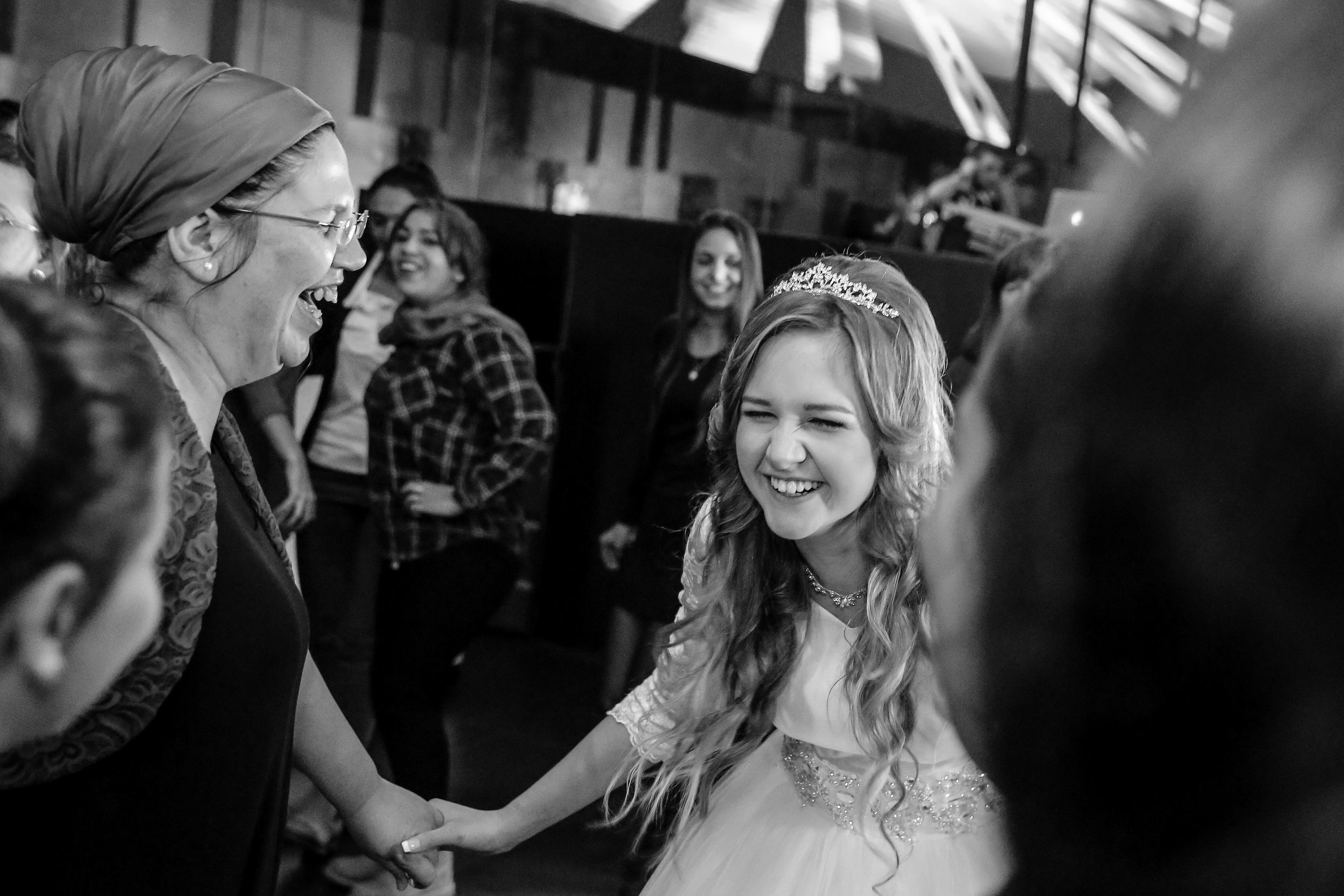 A black-and-white wedding photo capturing genuine laughter and joy: the happy bride laughs, holding hands with a woman at the wedding celebration. Both are smiling, sharing the joyous moment, with a blurred festive background.