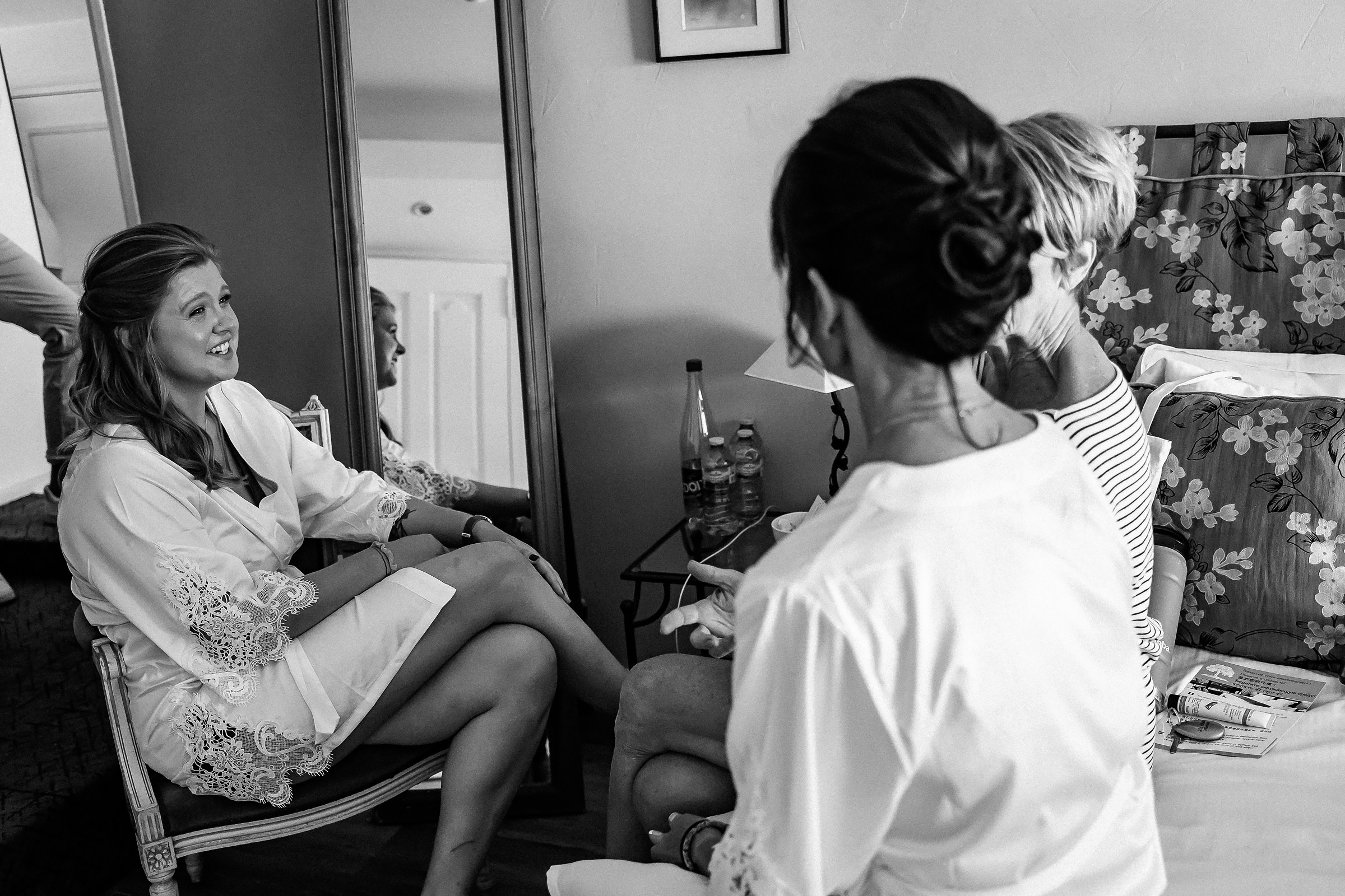 A cheerful bride chats with her friends while seated in a cozy bedroom in Provence, France, wearing a white lace robe.