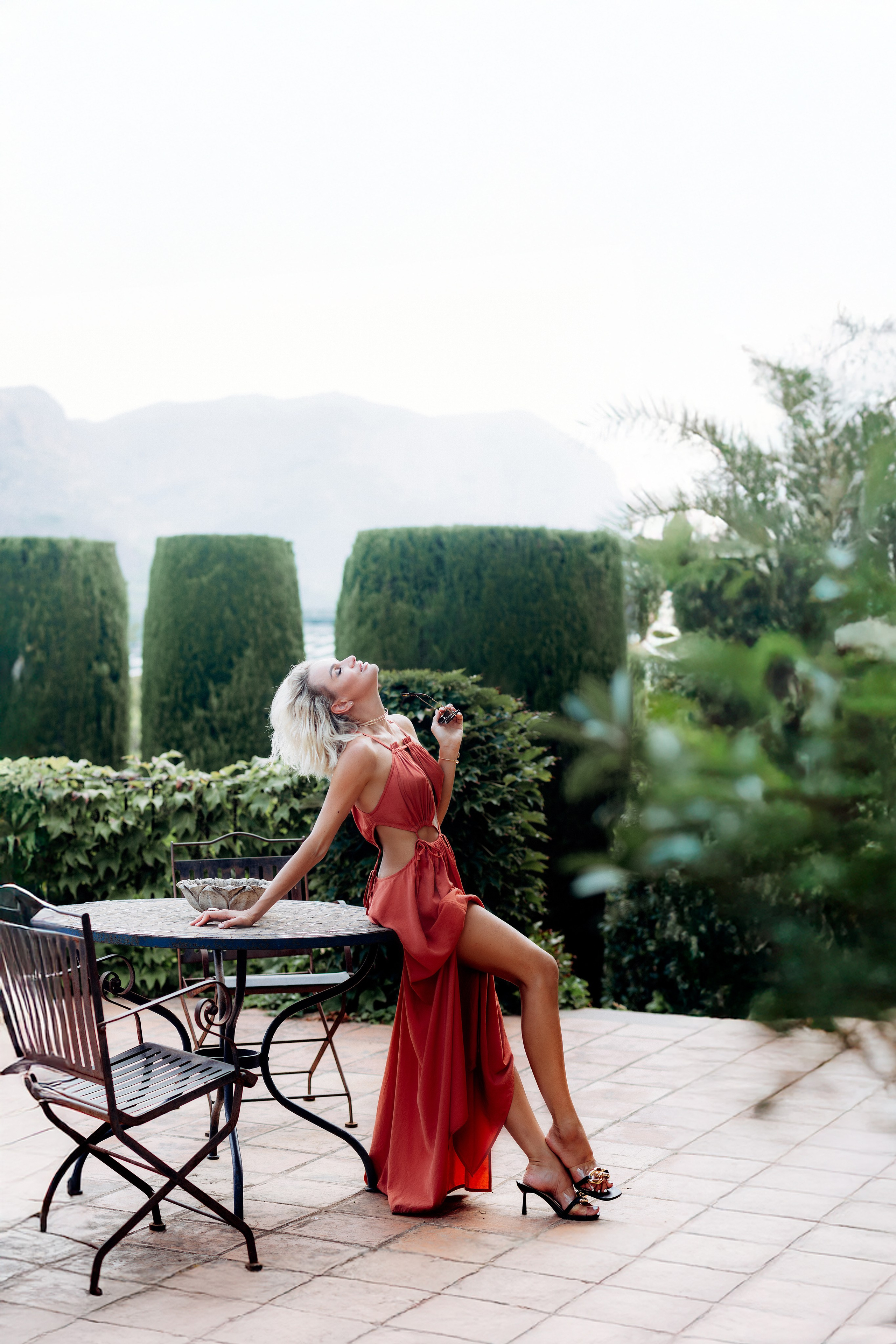 Элегантная портретная съёмка в Мадриде, Испания — стильная женщина в струящейся красной платье грациозно позирует на террасе, окружённой зеленью и мягким светом. Отличный вариант для тех, кто ищет гламурные и модные портреты в Мадриде и по всей Испании.