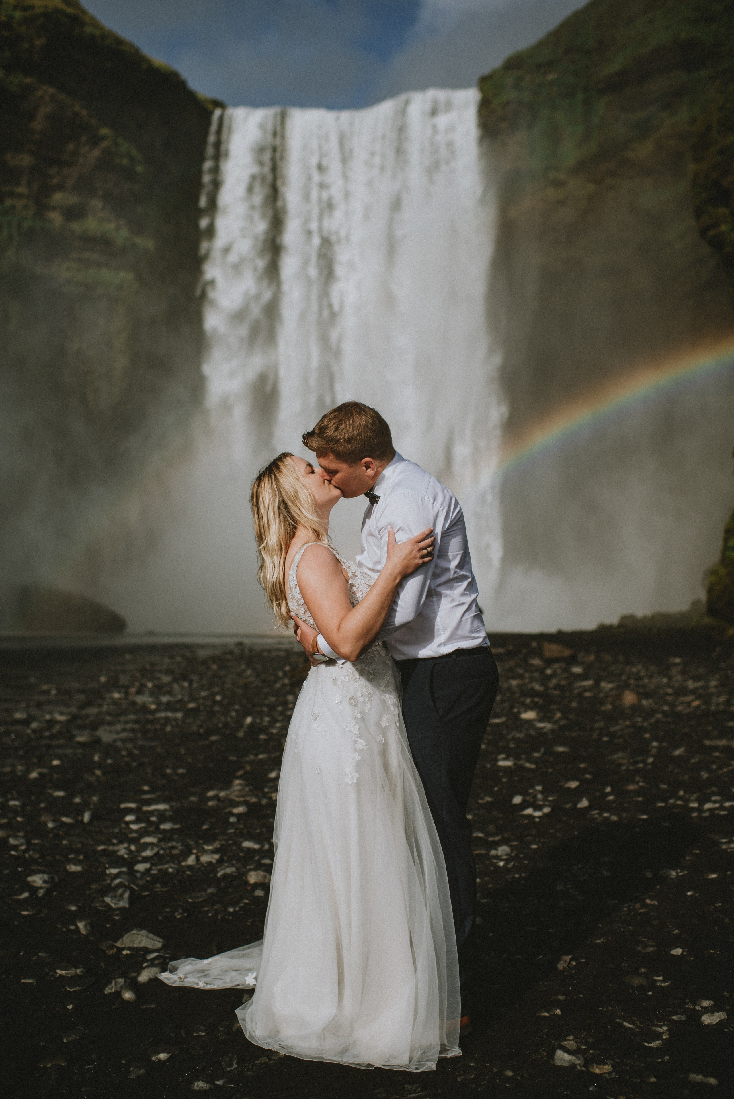 Bride and groom standing hand in hand in front of the majestic Skógafoss waterfall, mist rising around them.