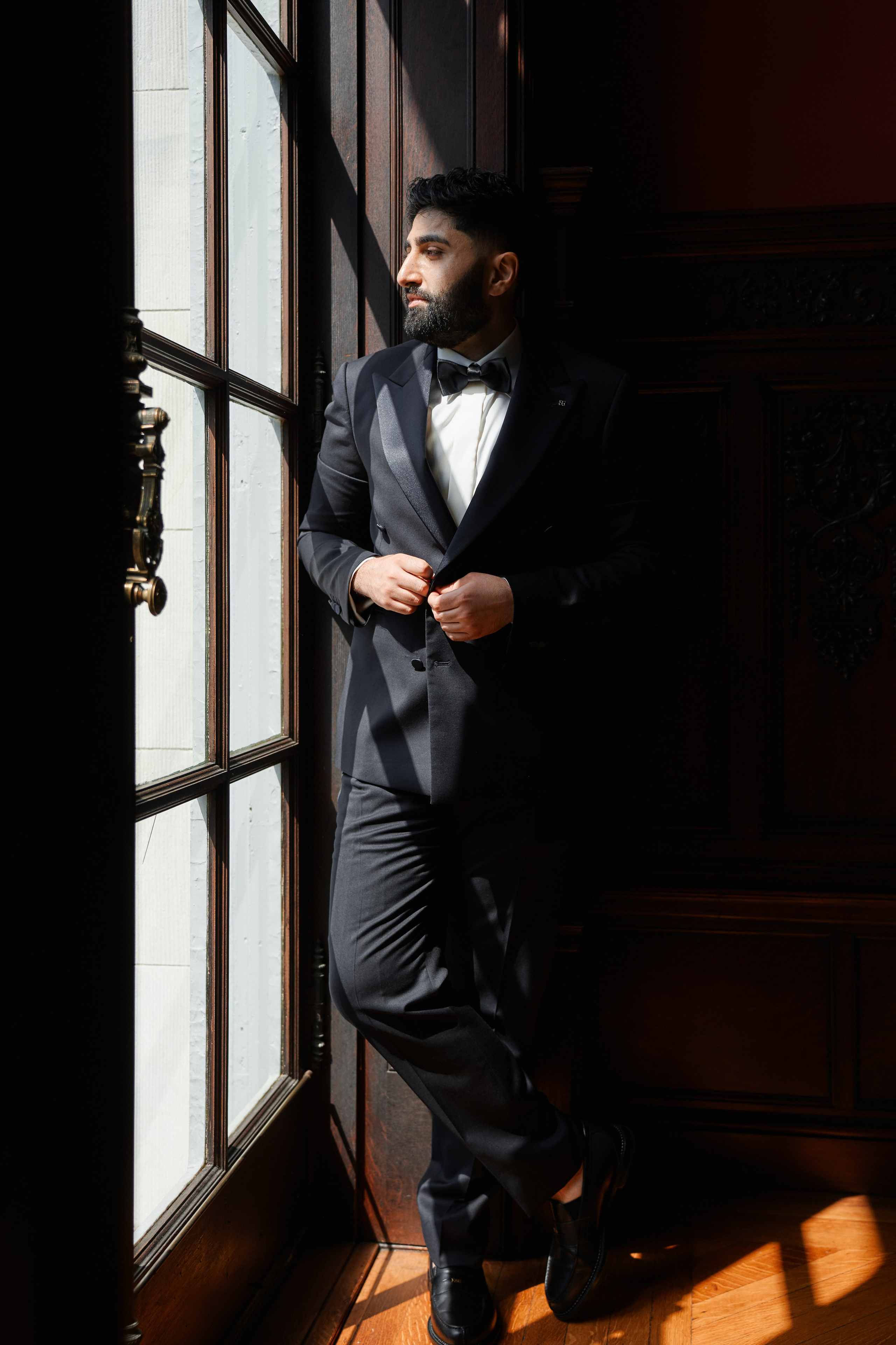 Stylish groom portrait in black tuxedo standing by window at luxury wedding venue