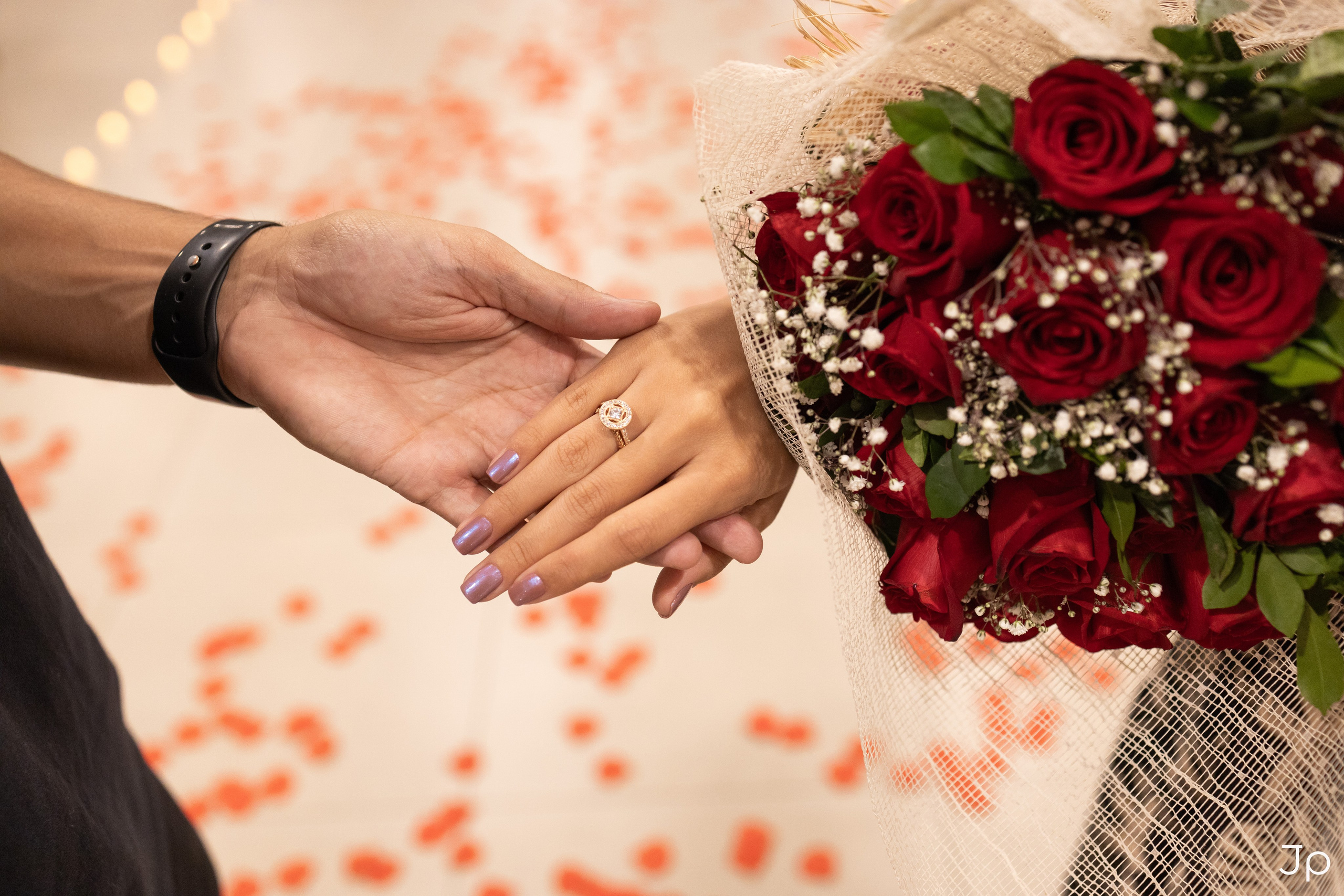 Close das mãos com alianças e buquê vermelho ao fundo.