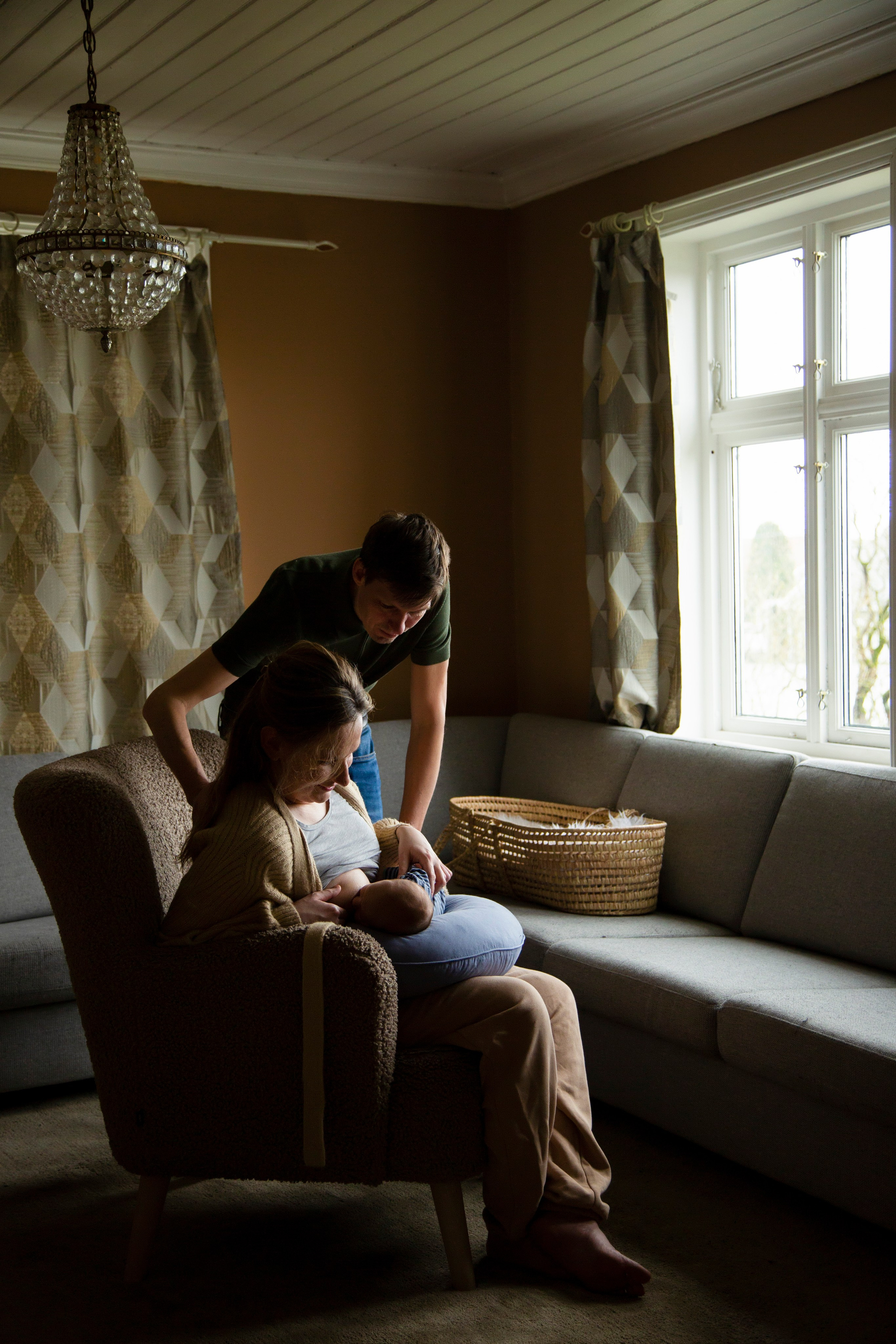 Mother sitting in a cozy armchair, cradling her newborn baby in her arms, with father standing behind her. Warm sunlight streams in through the window, creating a tender atmosphere