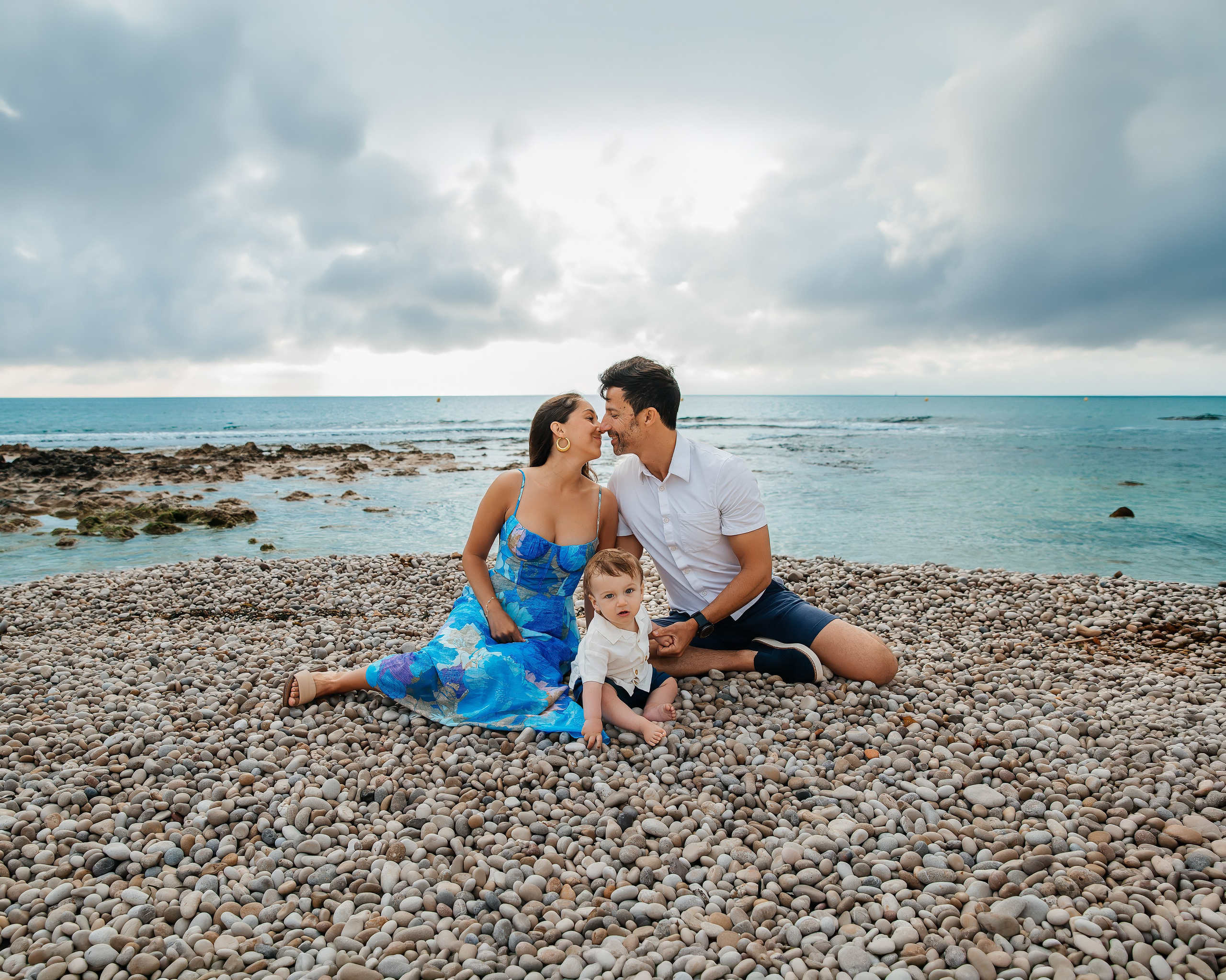 Sesión de fotos familiar en Valencia, España, con unos padres compartiendo un beso mientras están sentados en la playa junto a su pequeña hija con el mar Mediterráneo como telón de fondo. Un retrato familiar natural junto al mar que captura amor, unión y recuerdos inolvidables de sus vacaciones. Ideal para fotografía familiar, sesiones familiares en la playa, retratos familiares, fotografía de viajes y fotografía familiar de destino en Valencia y toda España.